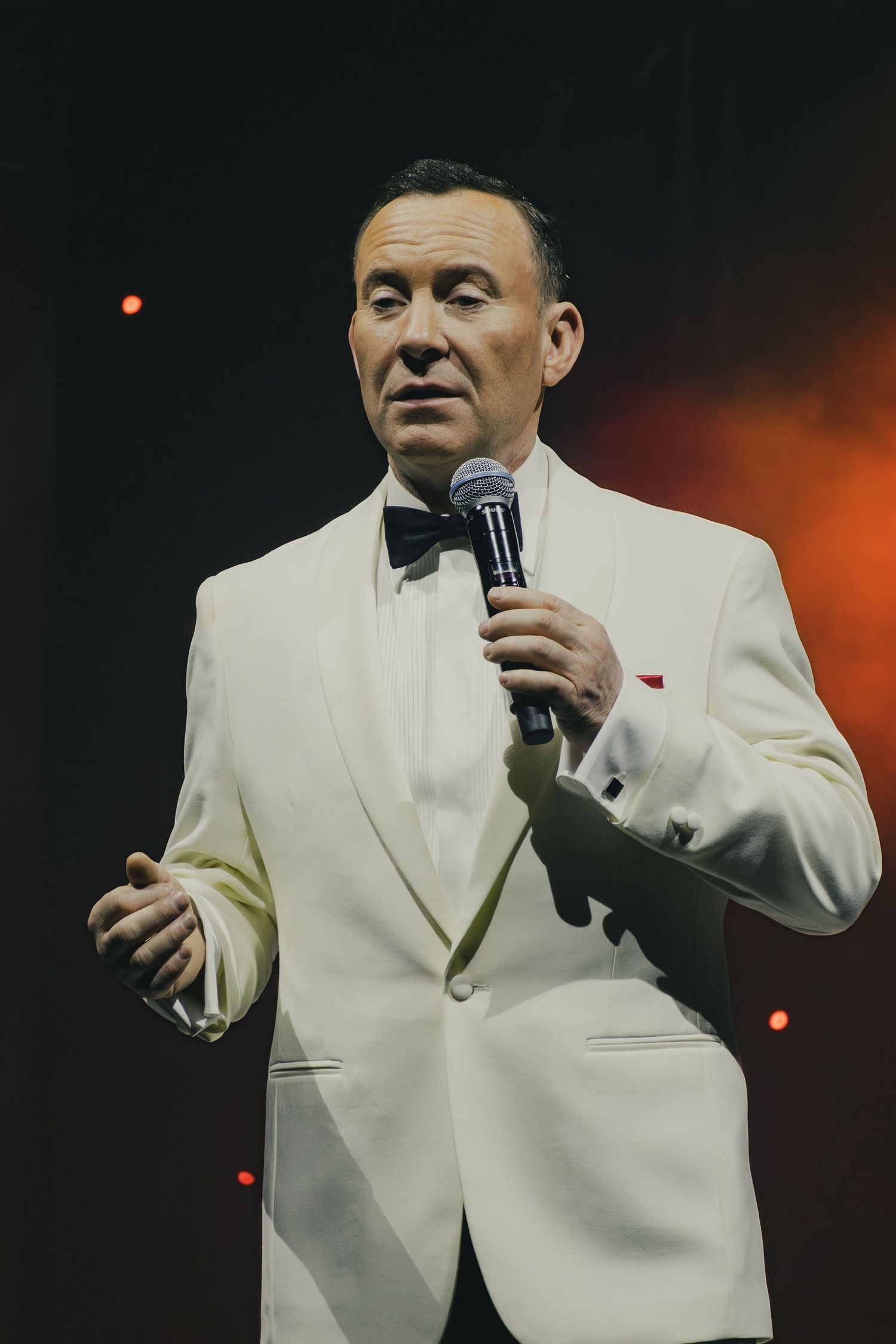 Performer in white tuxedo and bowtie holding microphone on darkened stage.