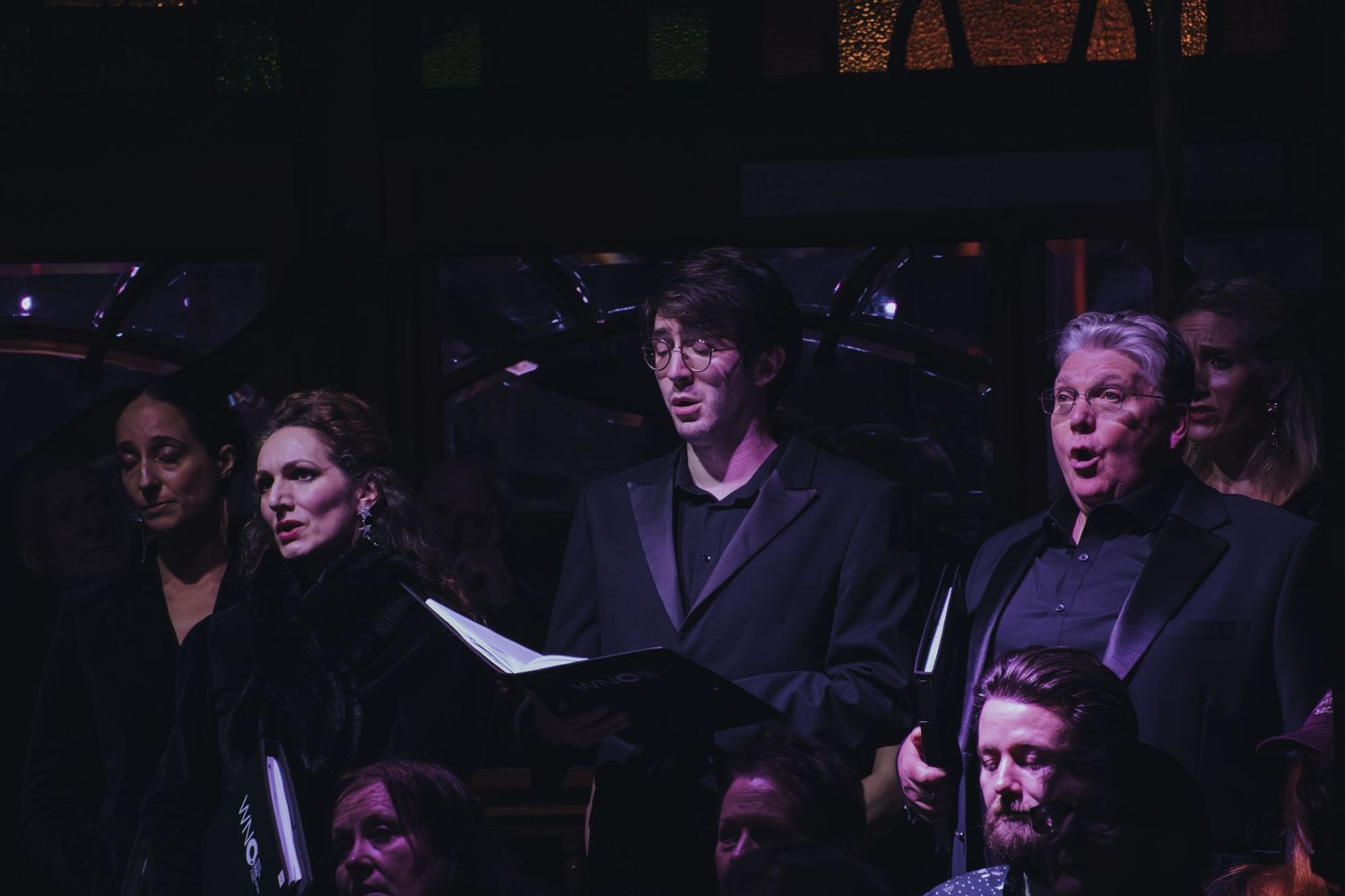 Choir members perform in formal attire under dramatic stage lighting.