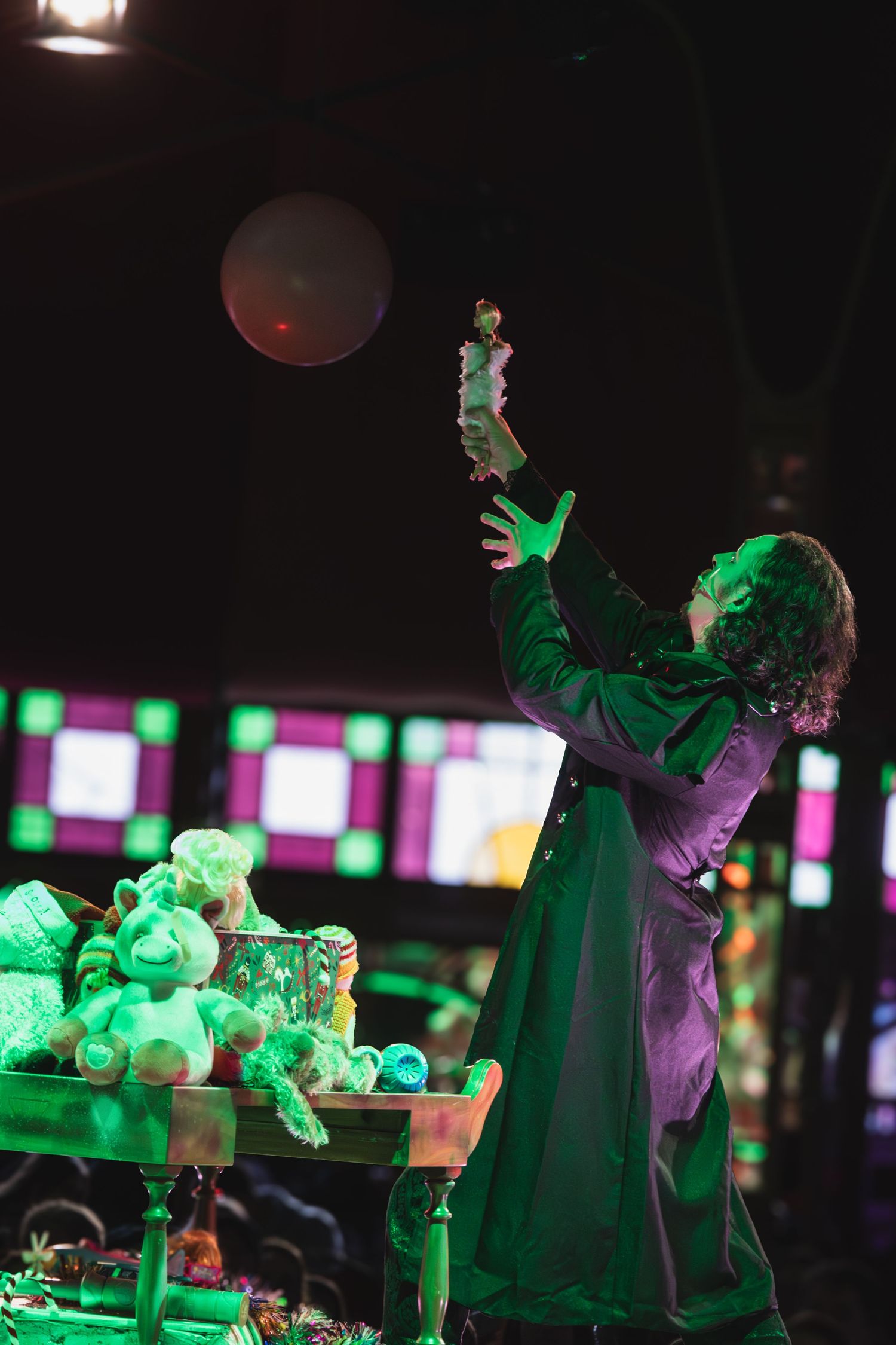 Stage performers in period costumes interact with props under green theatrical lighting.