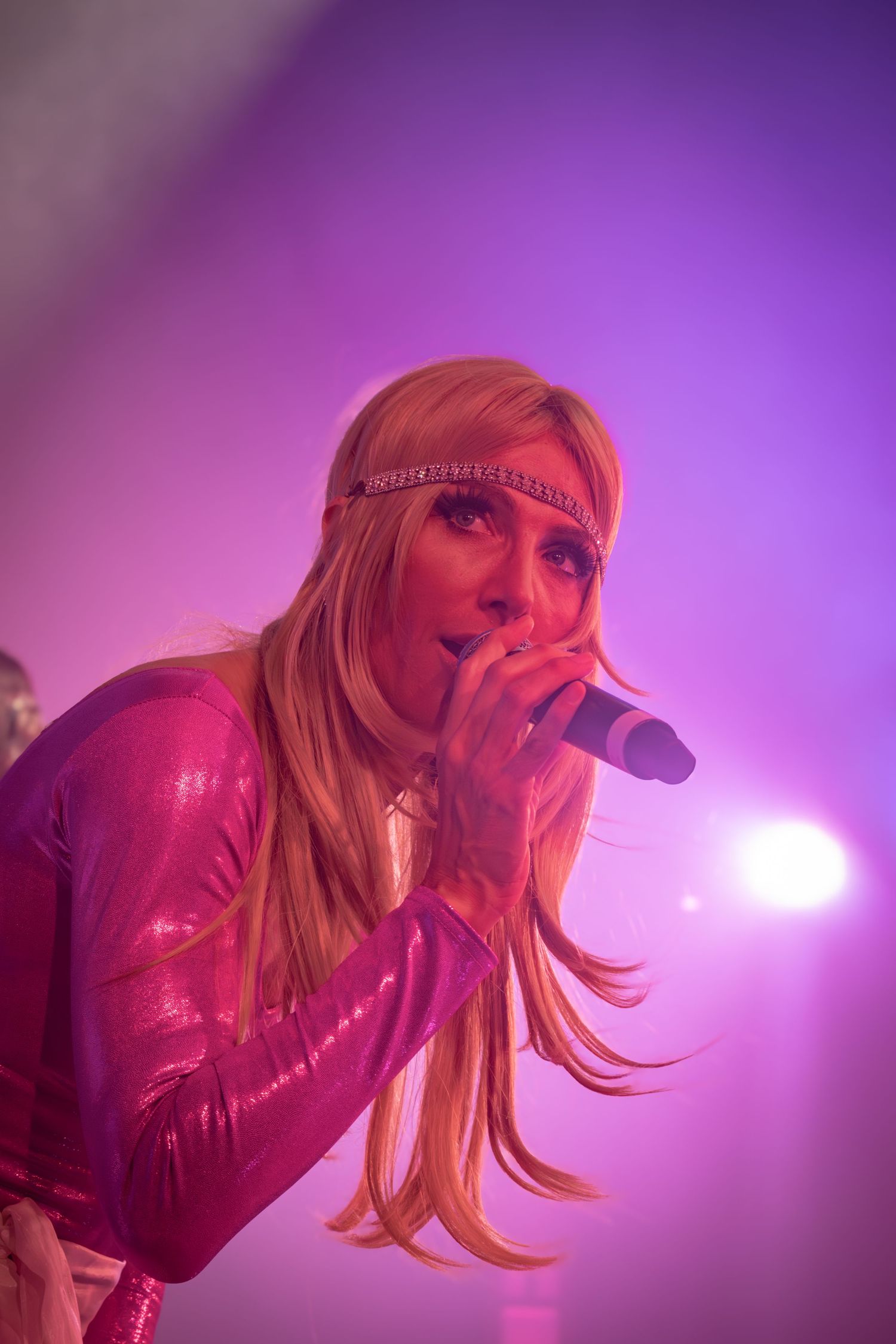 Singer in pink jacket leaning forward while performing under purple spotlights on stage.