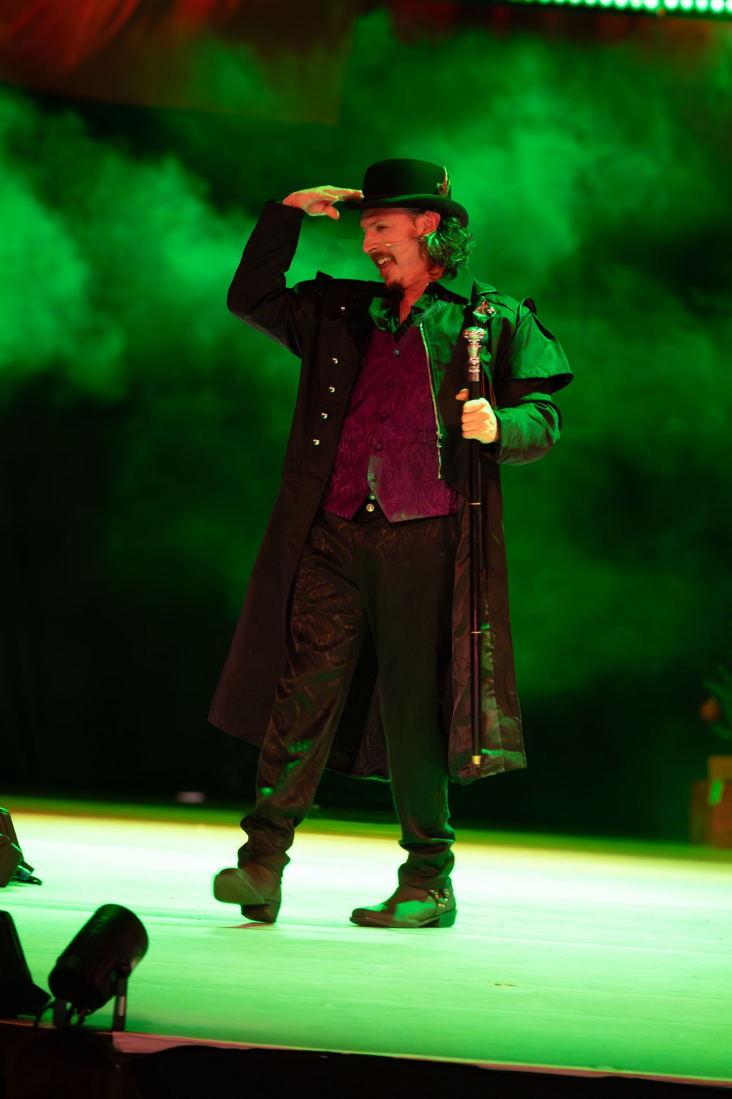 Stage performer in long coat and hat dancing in dramatic green lighting.