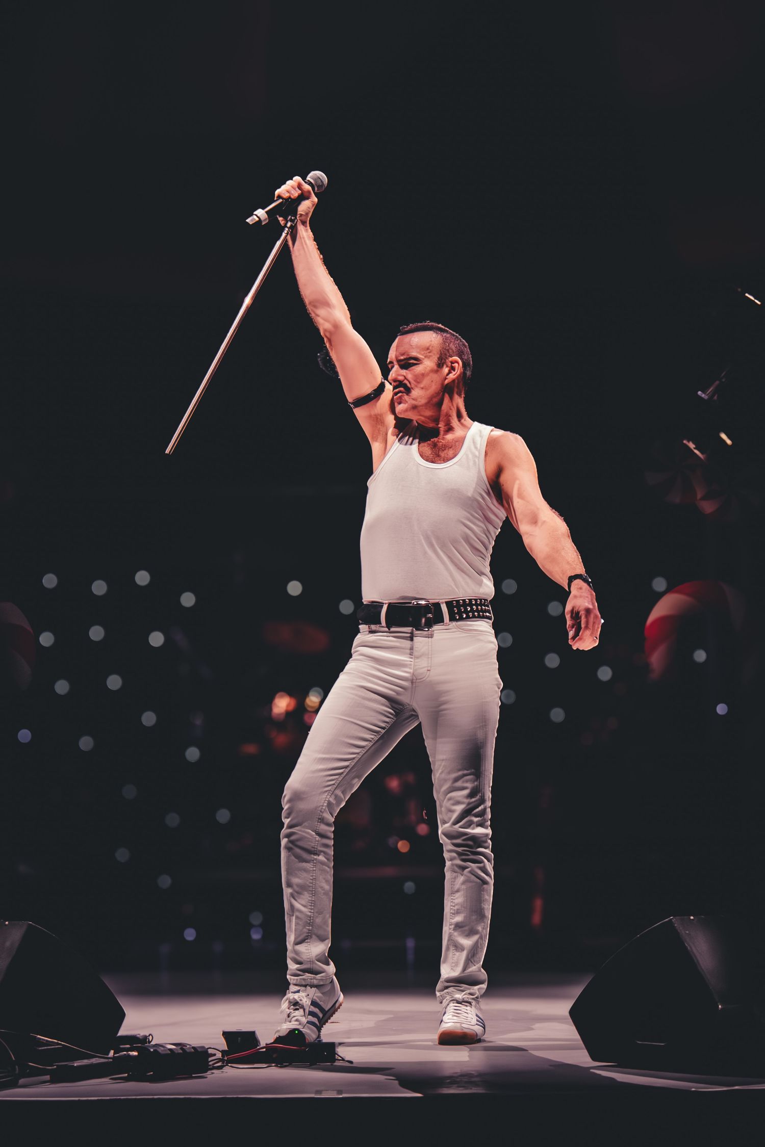 A performer in a white tank top and light pants strikes a dramatic pose on stage while holding a microphone up high.