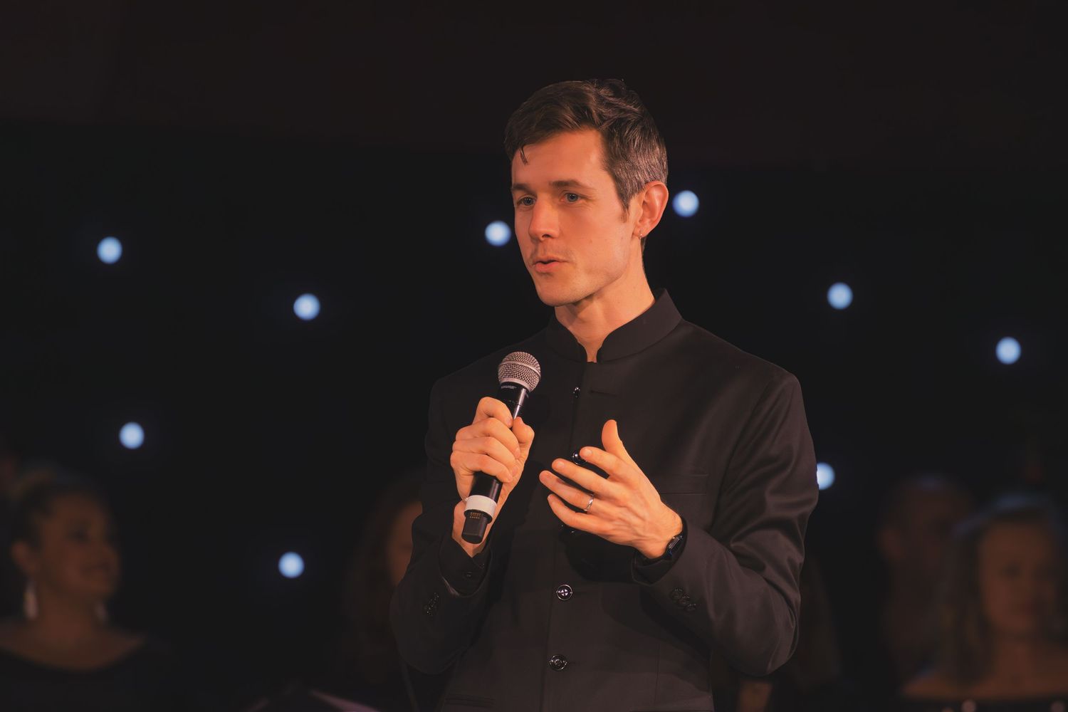 A performer in a dark suit speaks into a microphone on stage with blue lights illuminating the background.