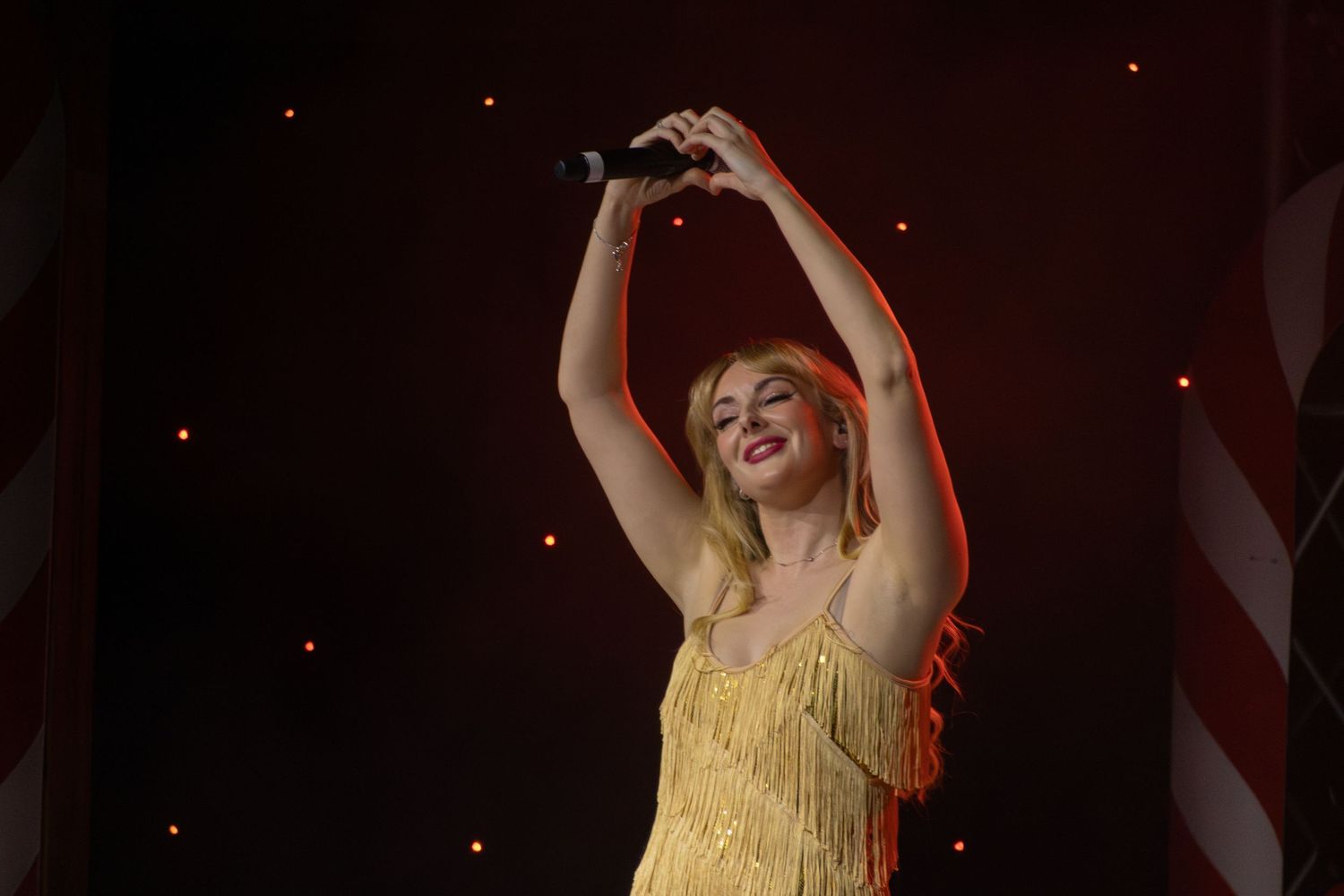 Performer in yellow dress raises arms during concert with dramatic lighting.