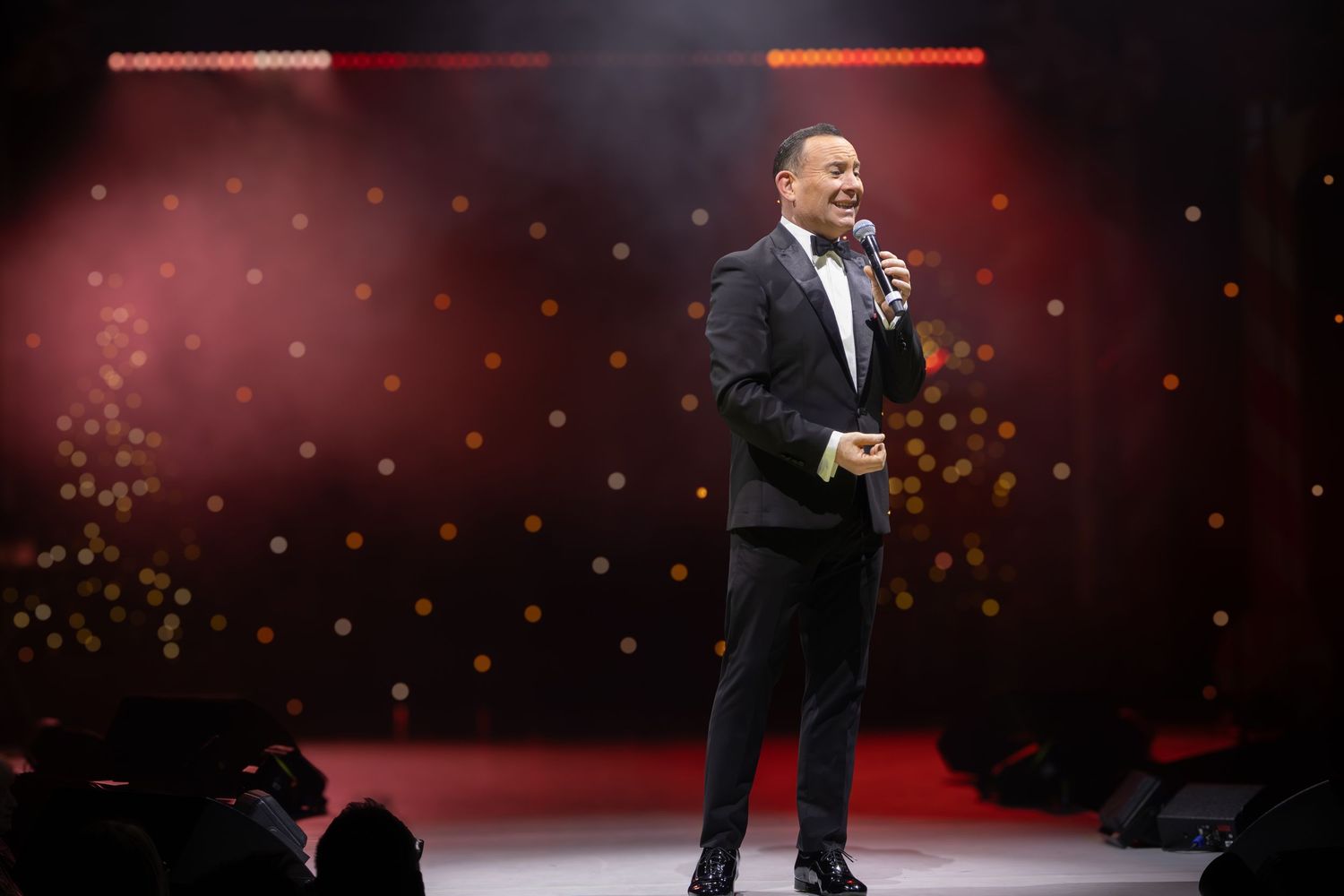 A performer in a black suit sings on stage with dramatic red lighting and bokeh effects.