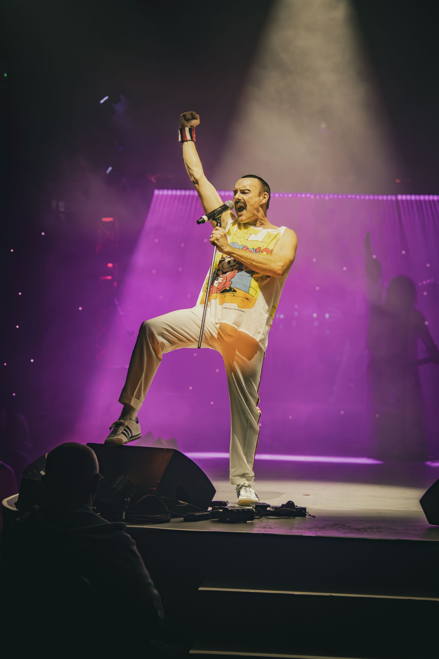 A performer in a white tank top and light pants strikes a dramatic pose on stage under purple lighting during a concert.