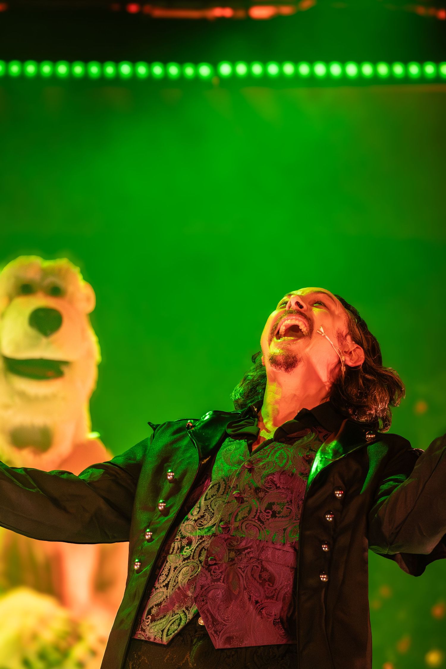 Theatrical performer gestures dramatically under green stage lighting.