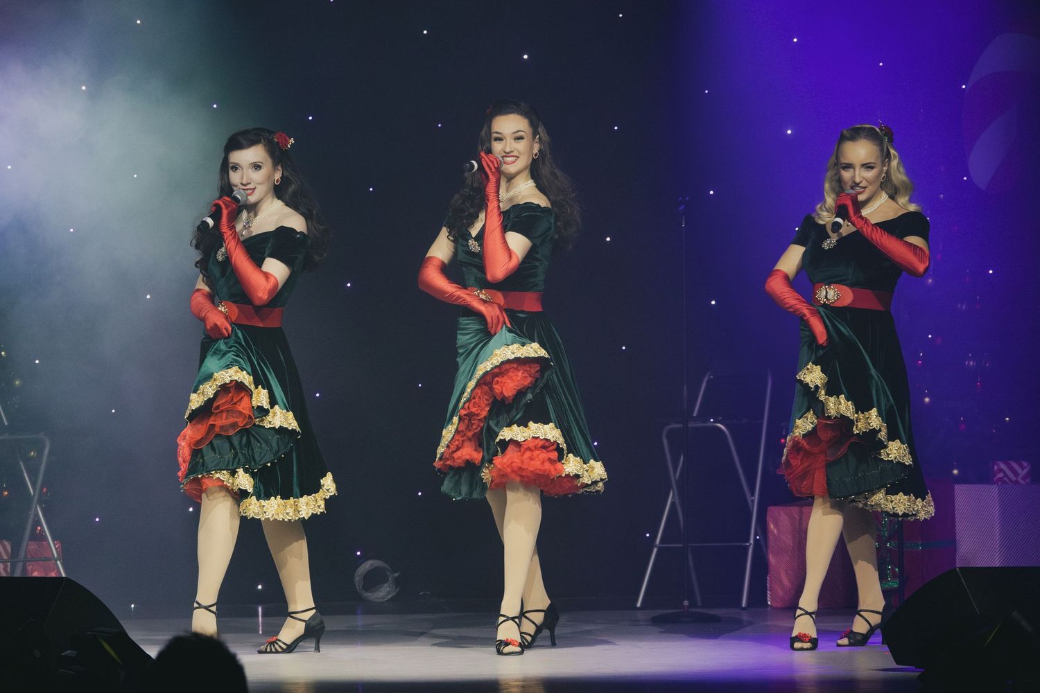 Three dancers perform in matching red and black dresses with ruffled skirts on a theatrical stage.