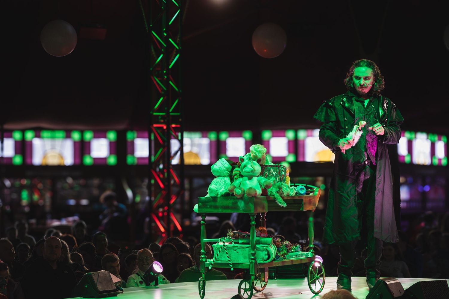 Stage performer in a green-lit theatrical scene with glowing screens in the background.