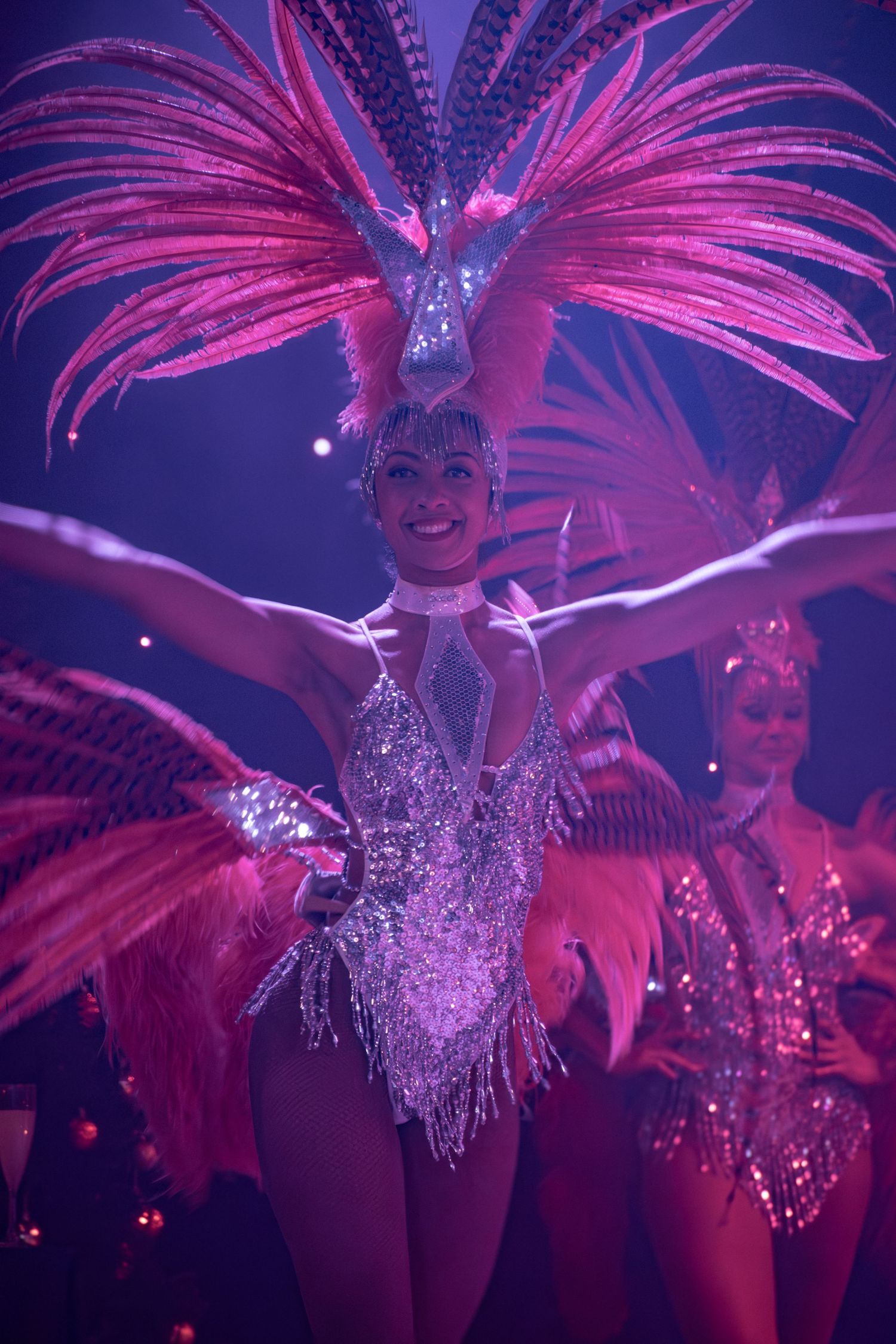 Showgirls in glittering costumes with pink feathered headdresses perform synchronized dance routine.