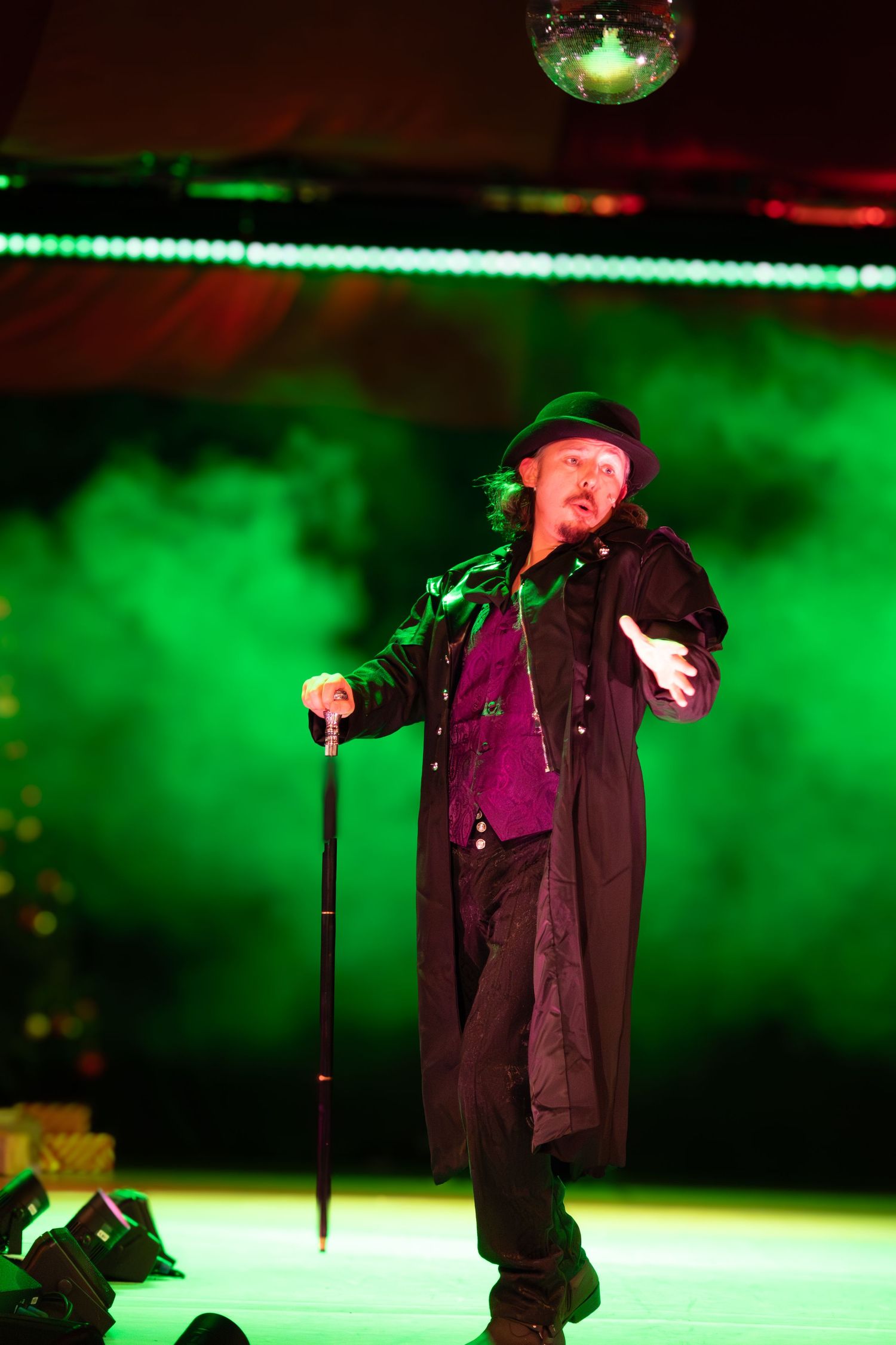 Performer in black coat and hat silhouetted against green fog on stage.