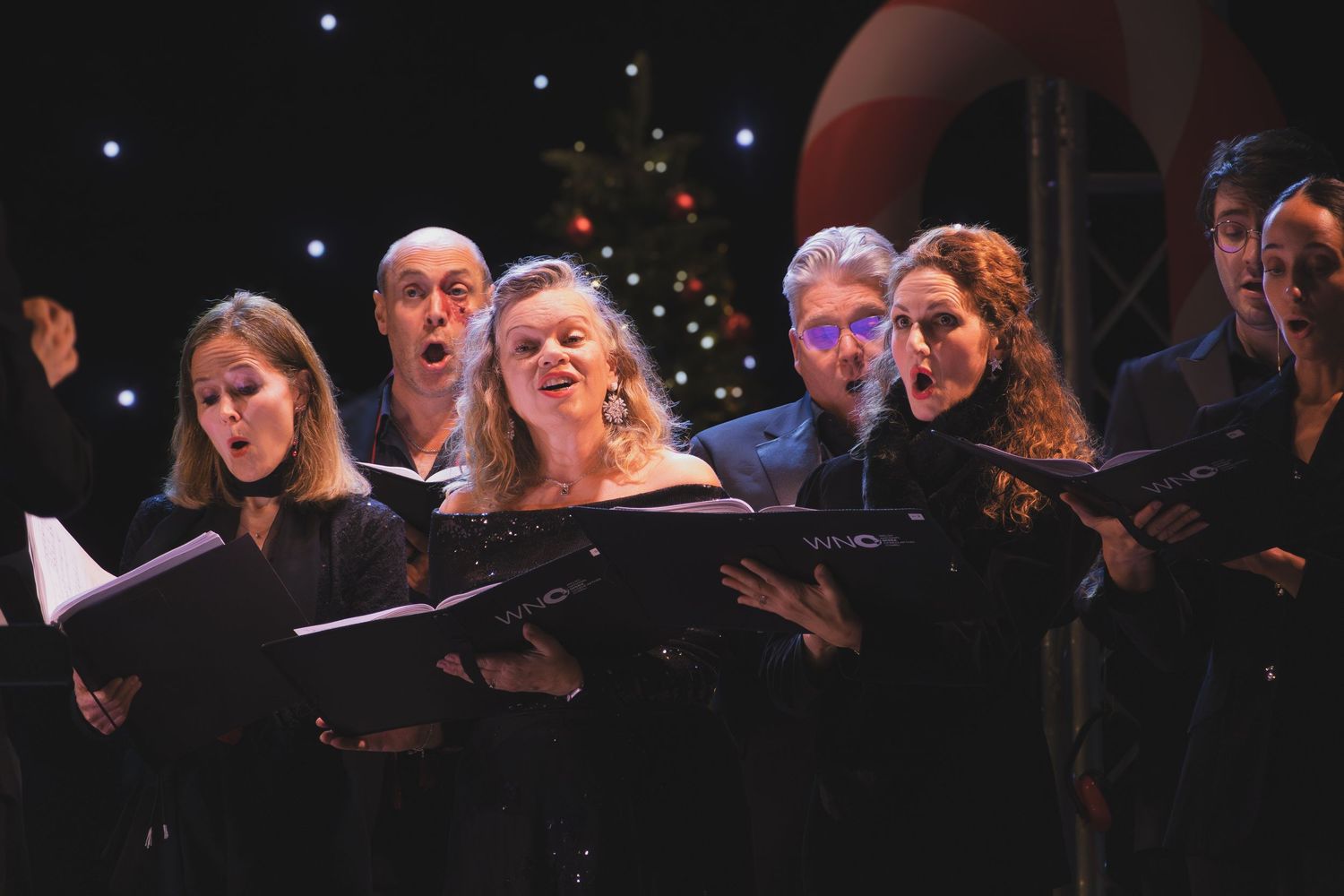 Choir members perform in formal black attire while holding sheet music under dramatic stage lighting.