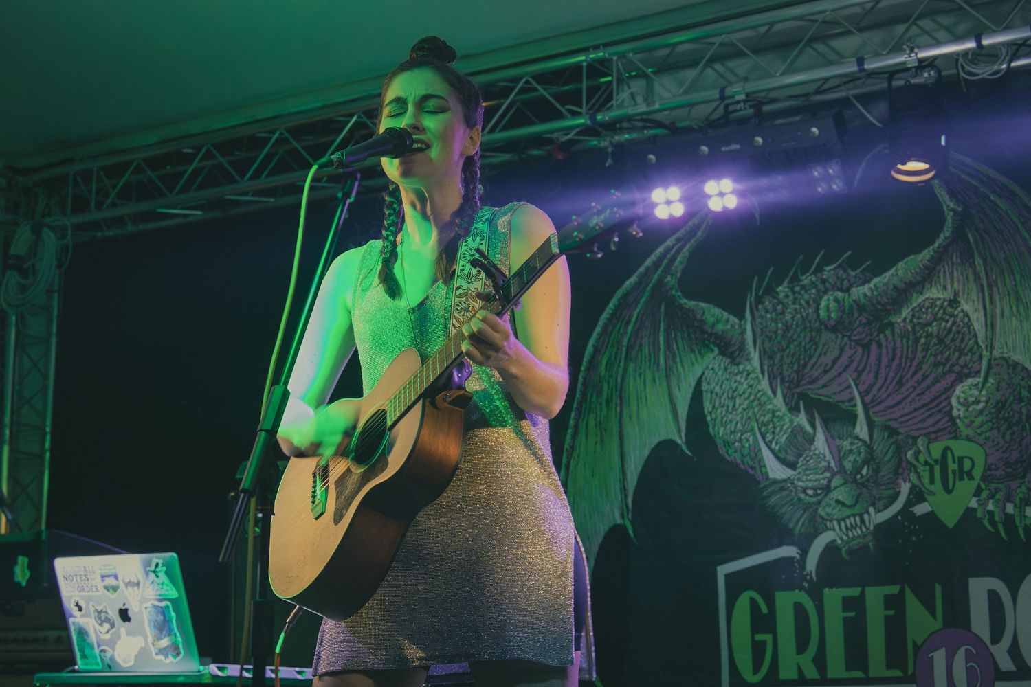 A performer plays acoustic guitar on stage with green lighting and a decorative backdrop.