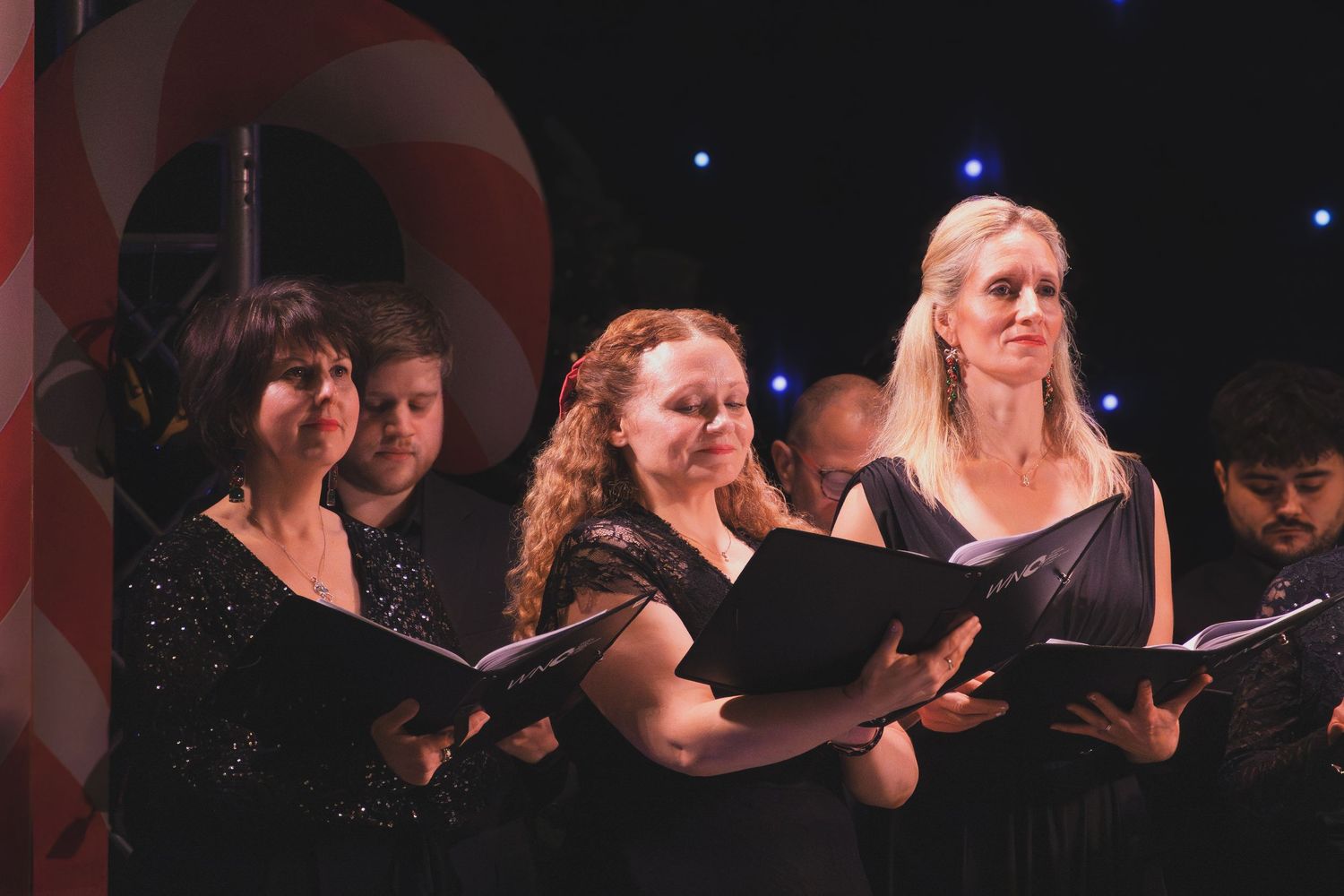 Performers singing in a choral ensemble against a dark background with stage lighting.