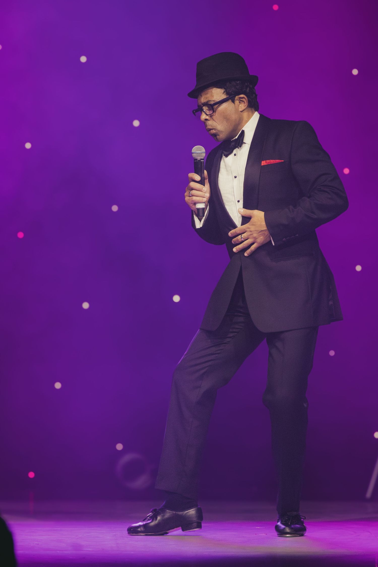 A performer in a fedora and tuxedo dances while singing on a purple-lit stage.