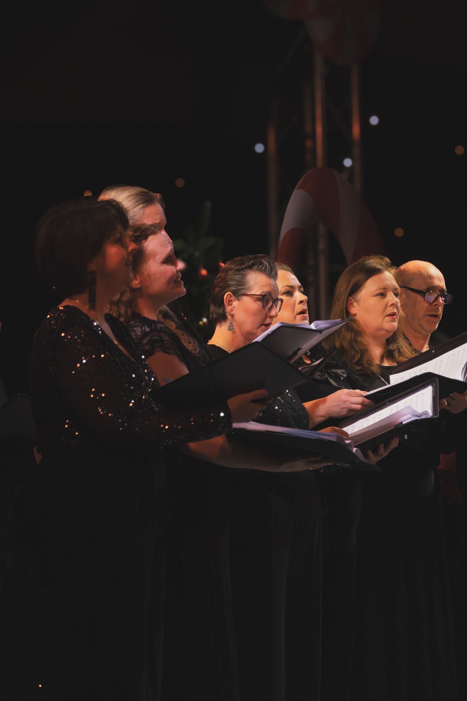 Choir members dressed in black hold music folders while performing on a dimly lit stage.