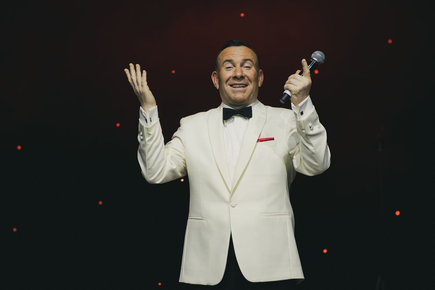 Performer in white tuxedo jacket gestures dramatically on dark stage with red lighting effects.