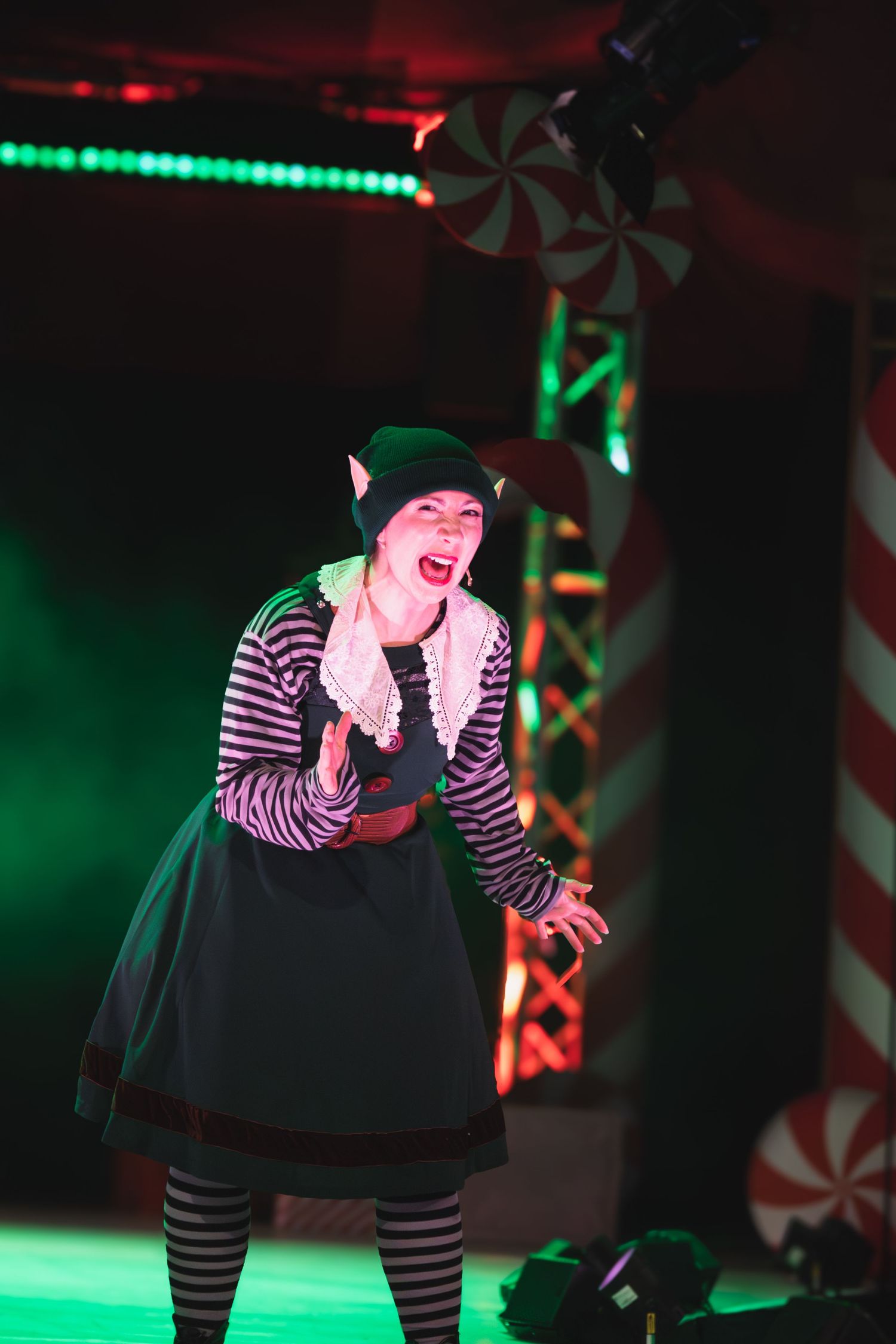 Theatrical elf character performs animated movements under red and green stage lighting.