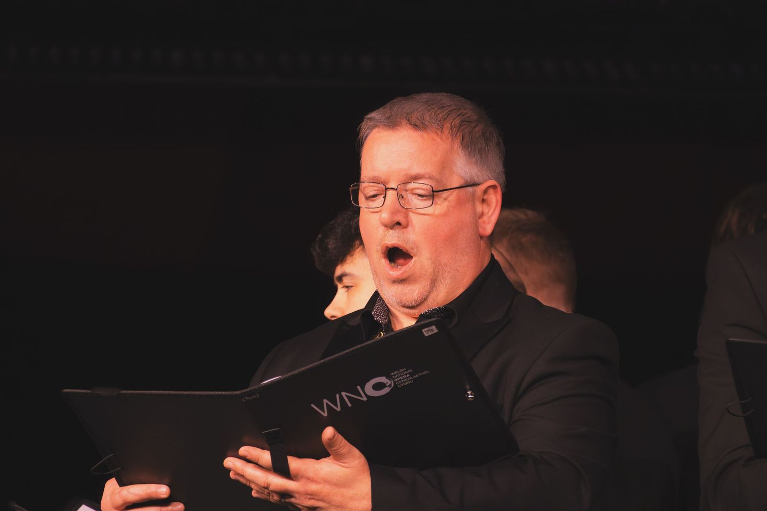 A choir member sings expressively while holding sheet music during an evening performance.