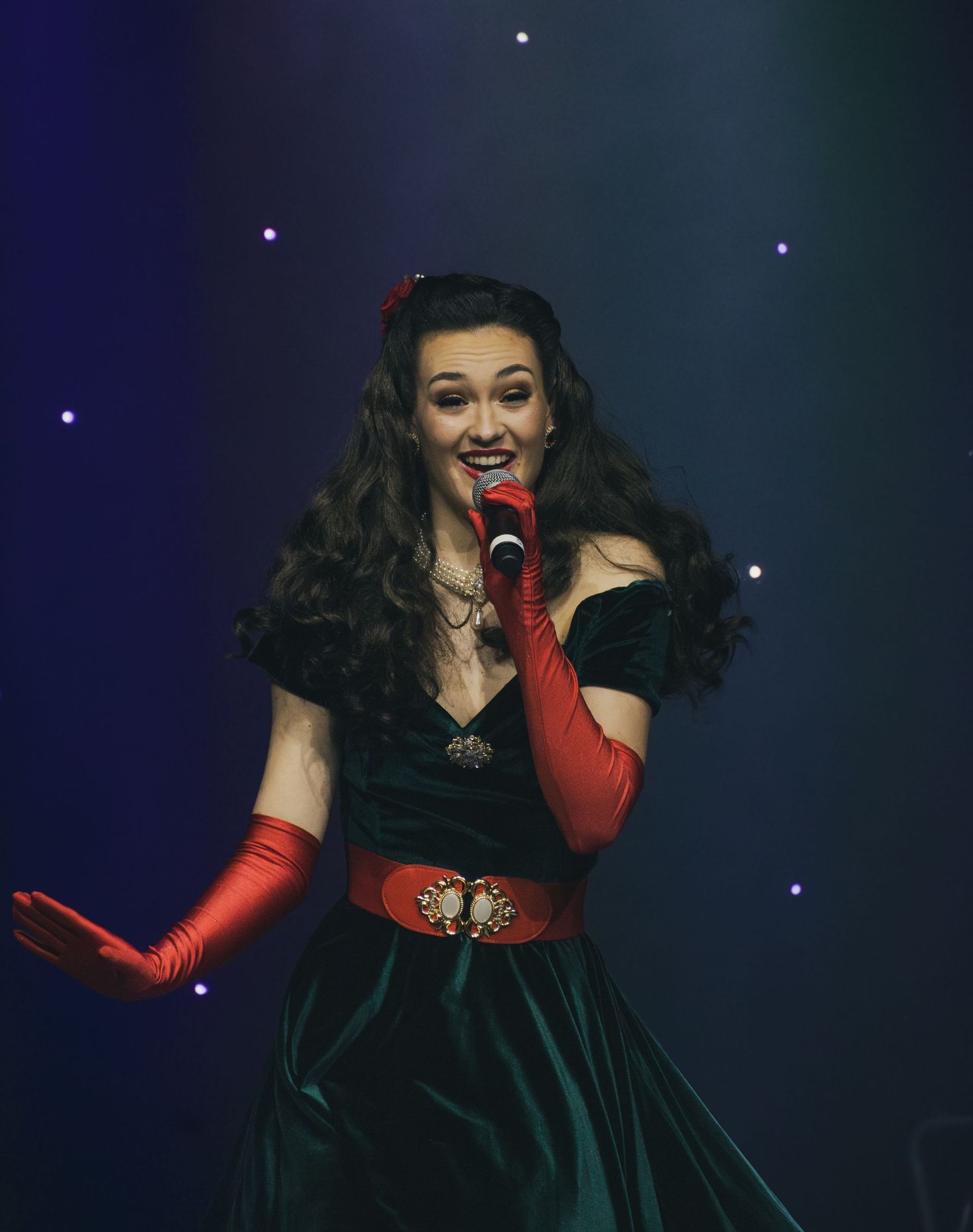 Performer in dark velvet dress with red gloves gestures dramatically on stage.