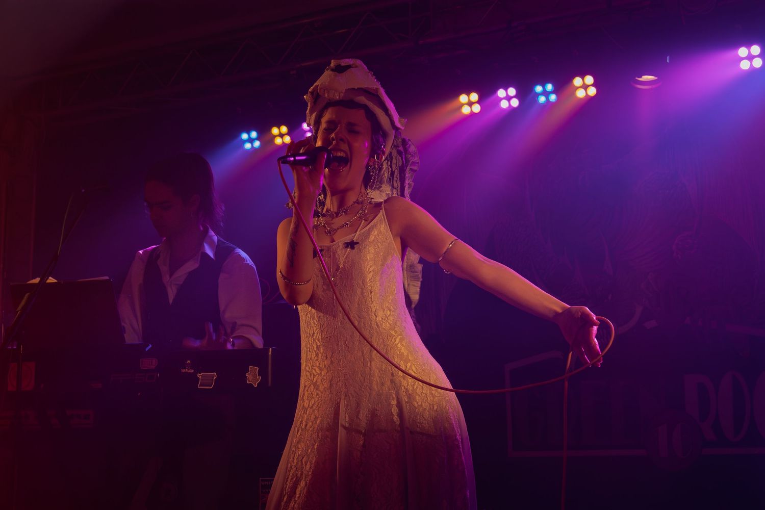 Concert performer in a white dress illuminated by vibrant purple stage lighting during a live show.