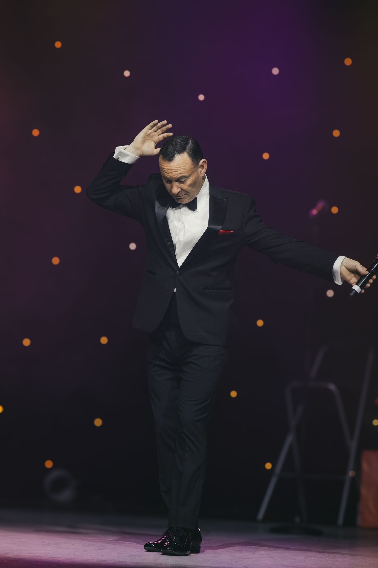 A performer in formal attire strikes a dramatic pose while holding a microphone on stage.