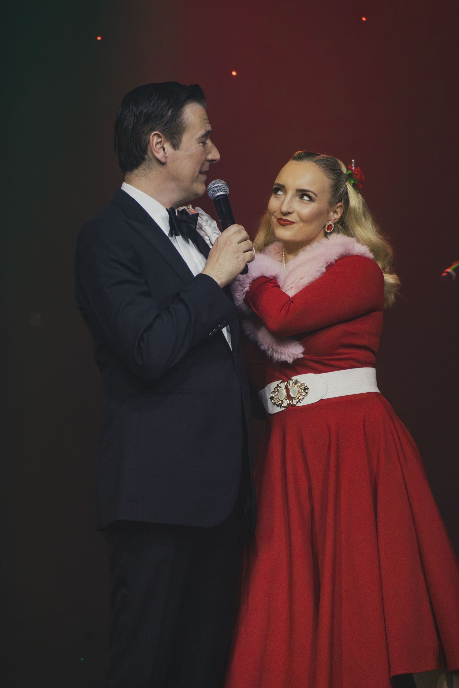 Two performers in red and black formal wear sing a duet on stage with dramatic lighting.