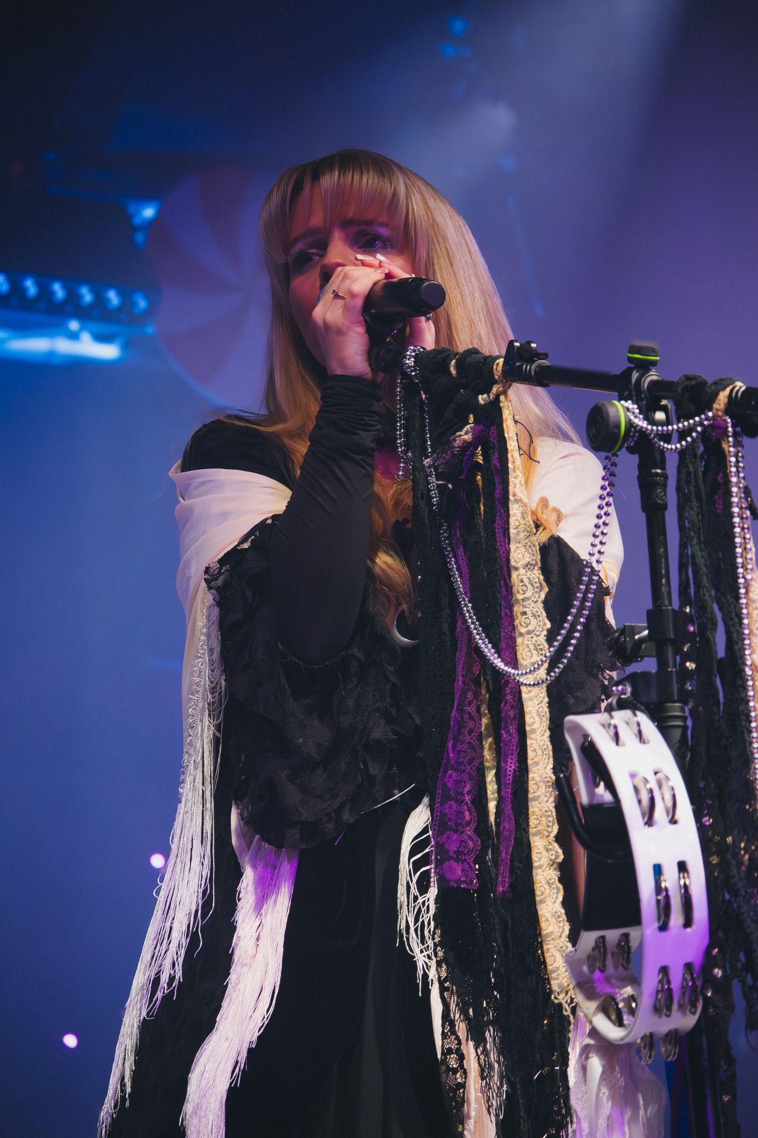 Performer in black and white outfit sings at decorated microphone stand under blue lights.