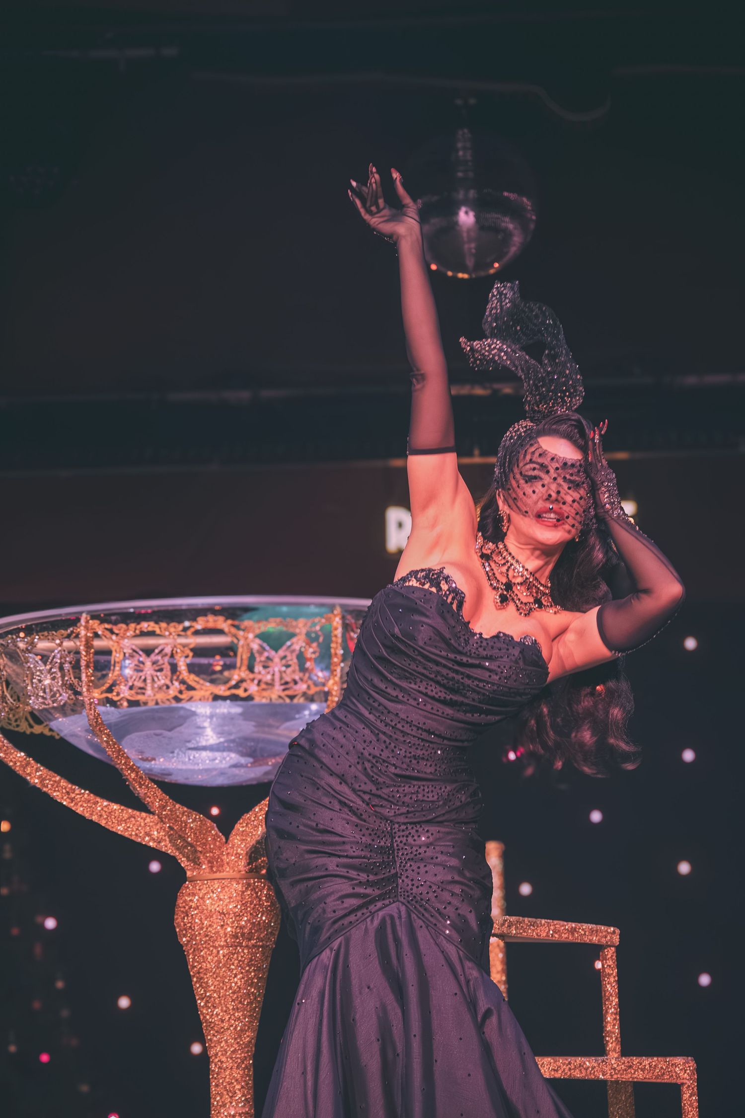 Burlesque performer in black dress dances dramatically next to golden decorative stage props.
