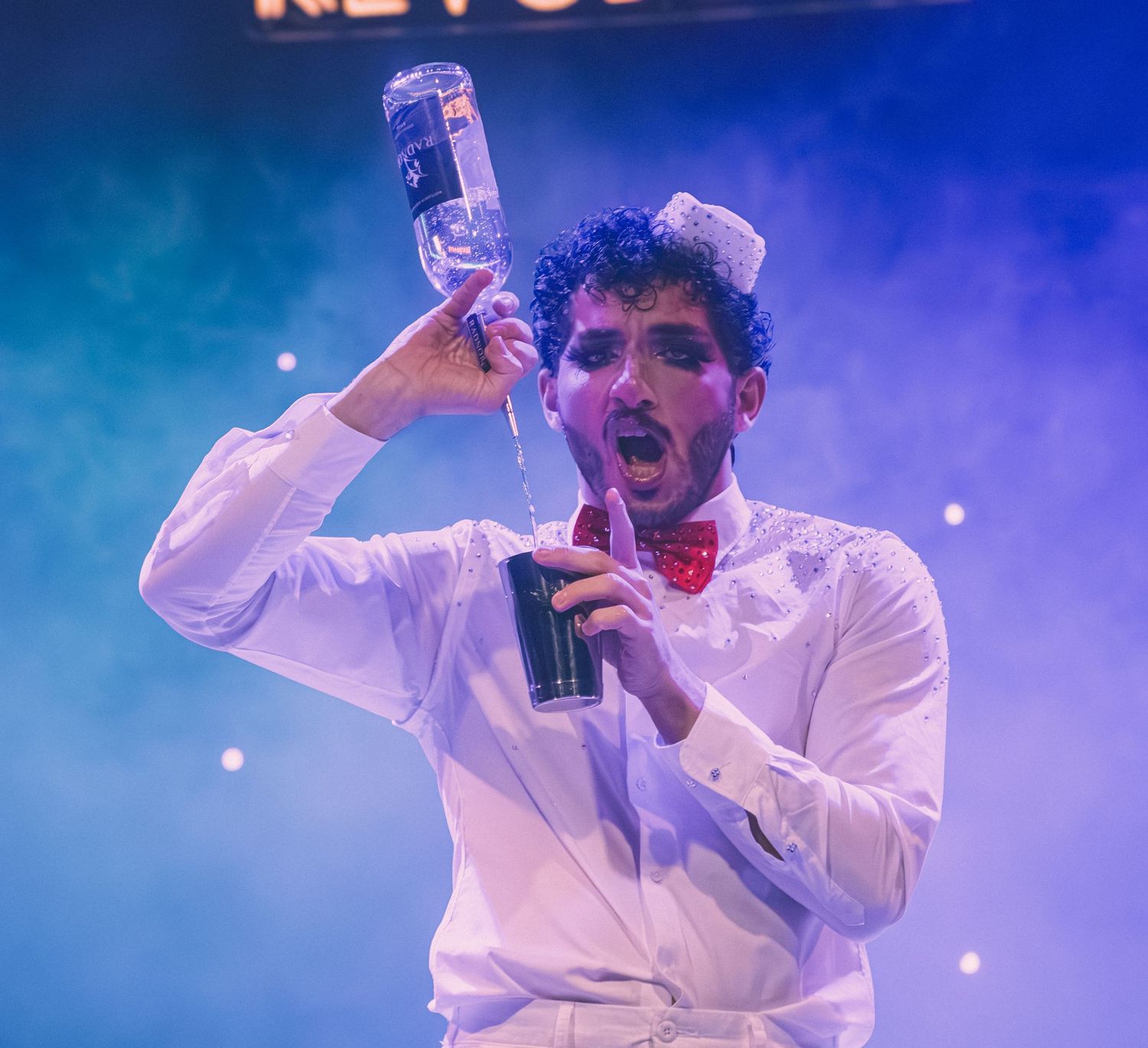 A performer in white attire sings dramatically while holding a bottle on stage.