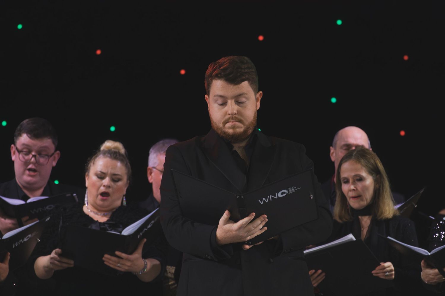 Choir members in formal black attire perform under colored stage lights while reading from music books.