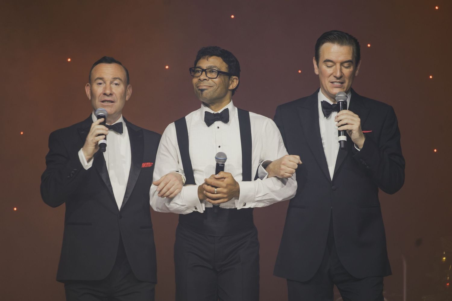 Performers in tuxedos sing and gesture during a stage performance.