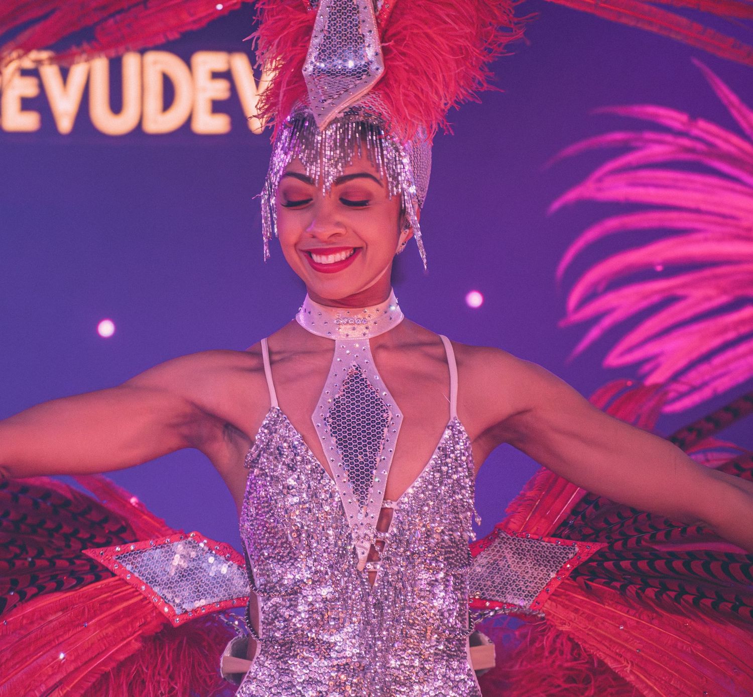 Burlesque performer in sparkling silver costume with red feathers performs on stage under purple lighting.