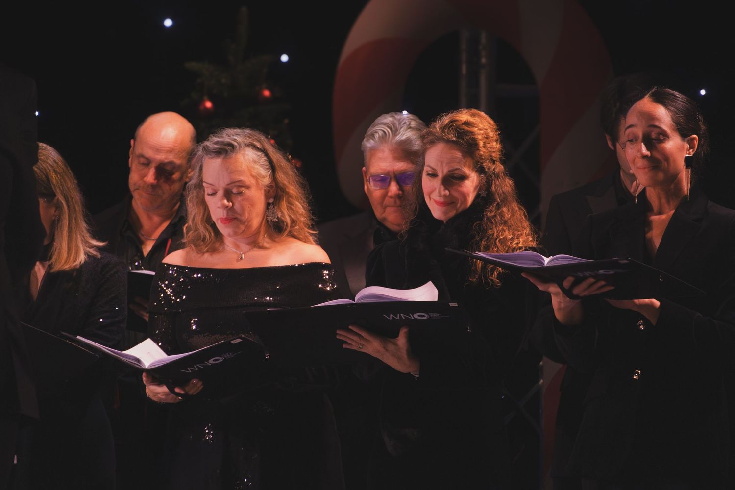 Choir singers dressed in black perform while reading from music books on a darkened stage.