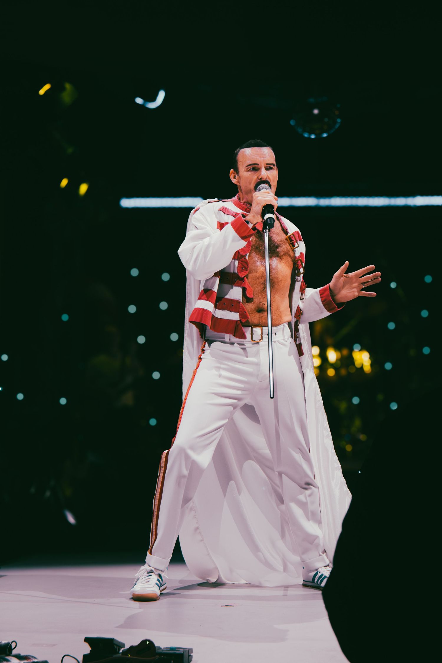 A performer in a white jumpsuit kneels dramatically on stage during a concert performance.