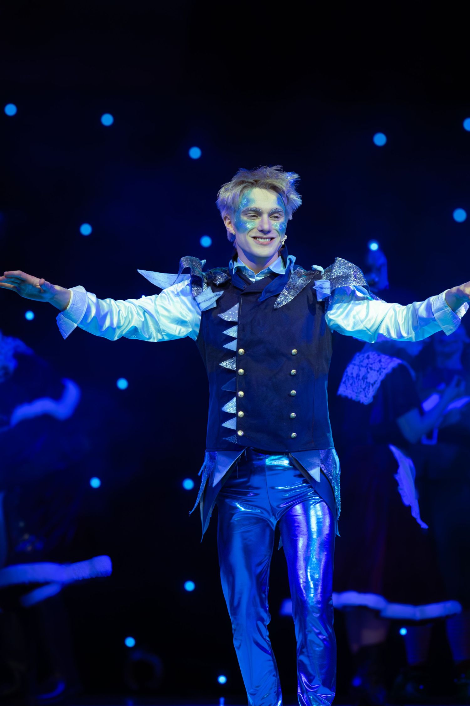 Performer in blue costume spreads arms wide during dramatic stage moment under blue lighting.