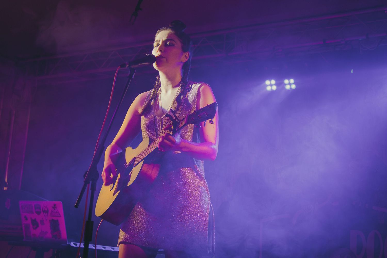 Musicians perform on stage with purple lighting and fog effects creating a dramatic concert atmosphere.
