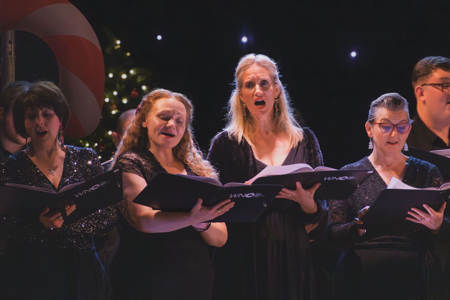 Performers in black attire sing together during a choral concert with colorful lights in background.