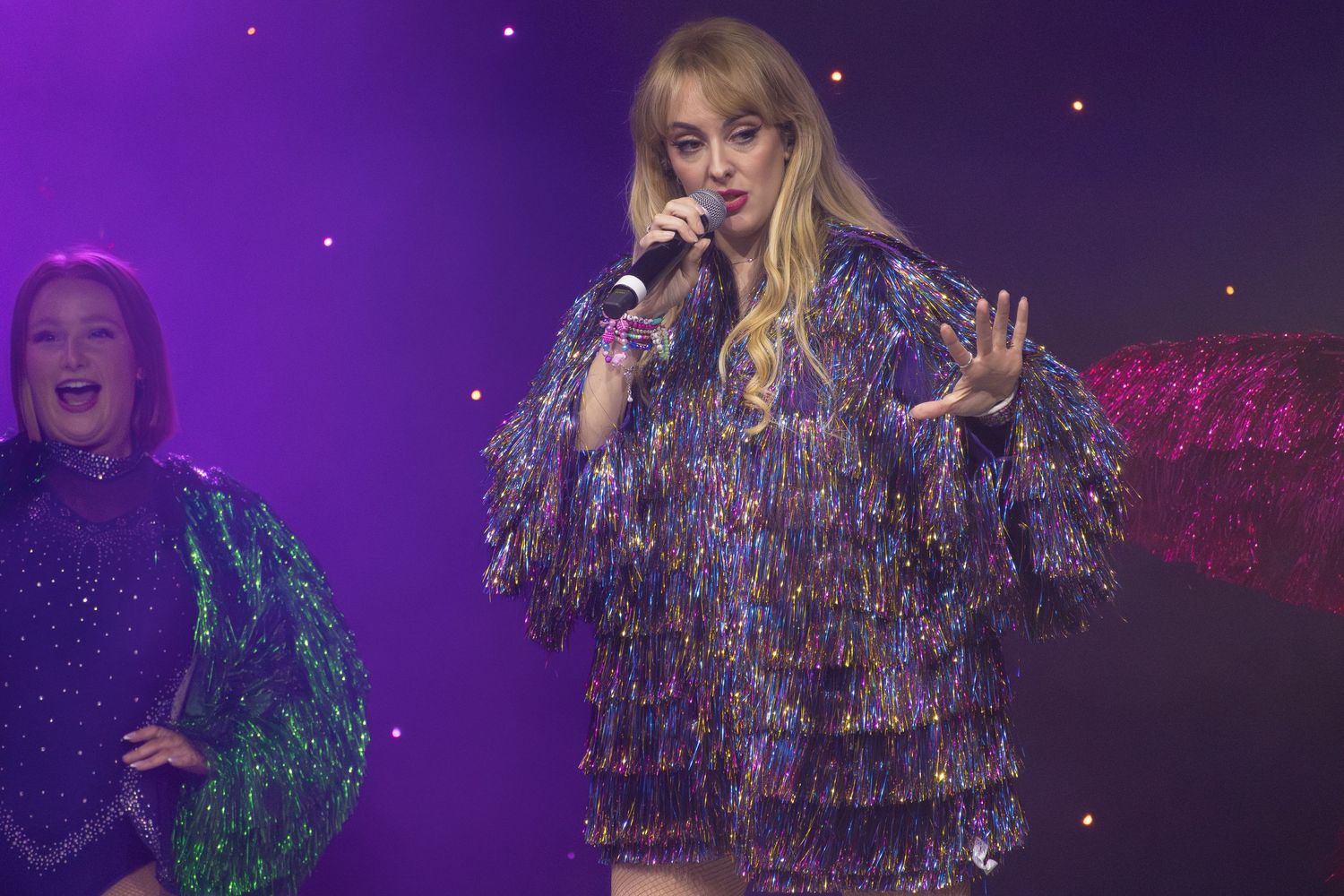 Concert performer in a sparkly dress and boots dances energetically on a purple-lit stage.