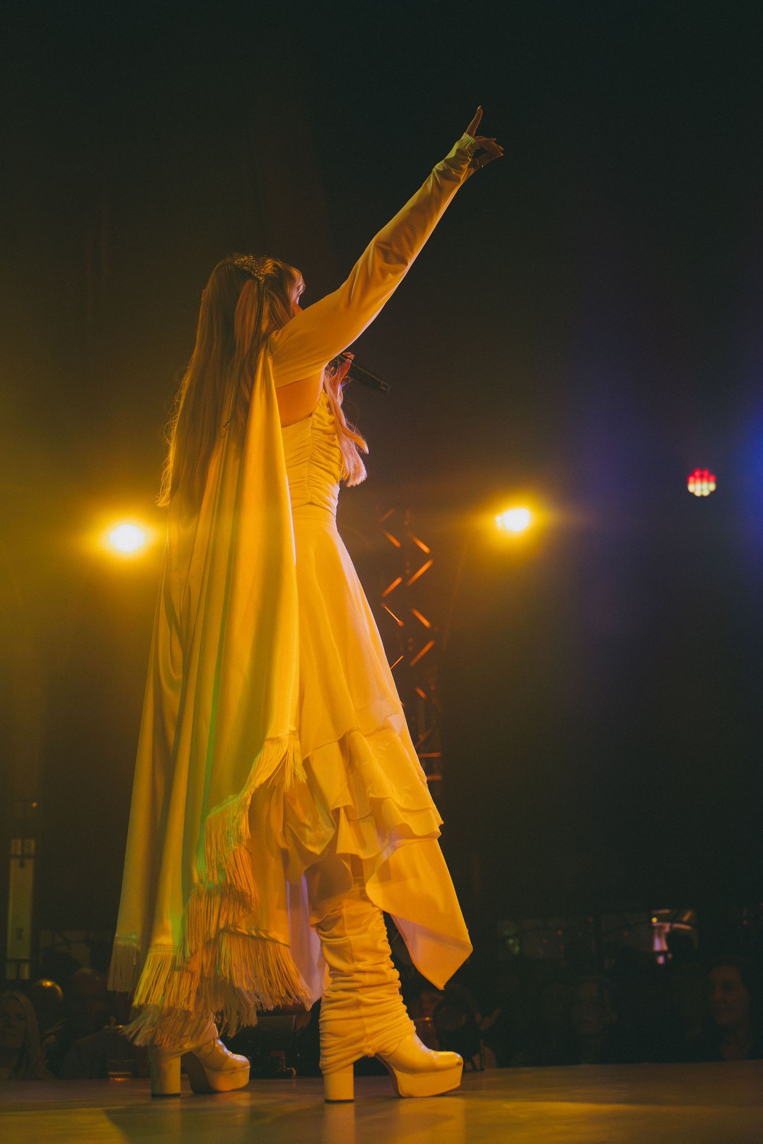 Performer in flowing white costume moves dramatically on darkened stage with amber lighting.