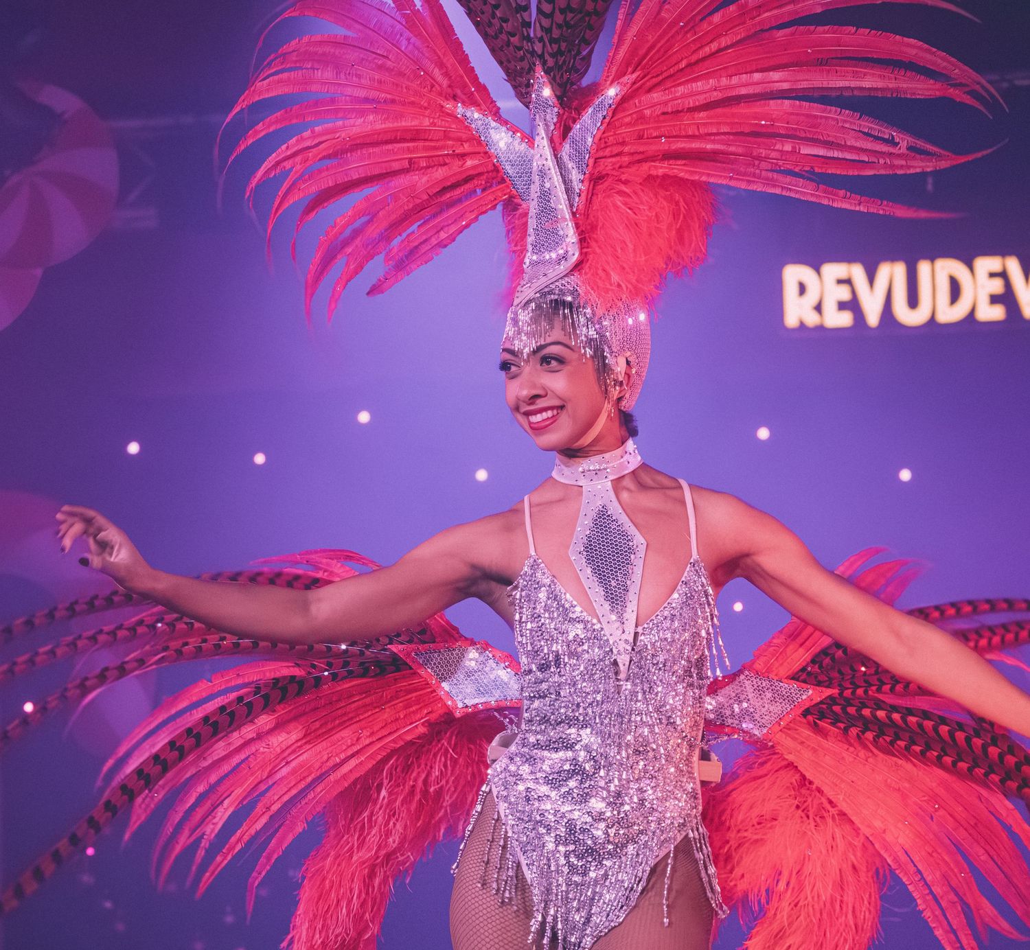 A showgirl performer in a sparkly costume with vibrant pink feathers dances at Revudeville.