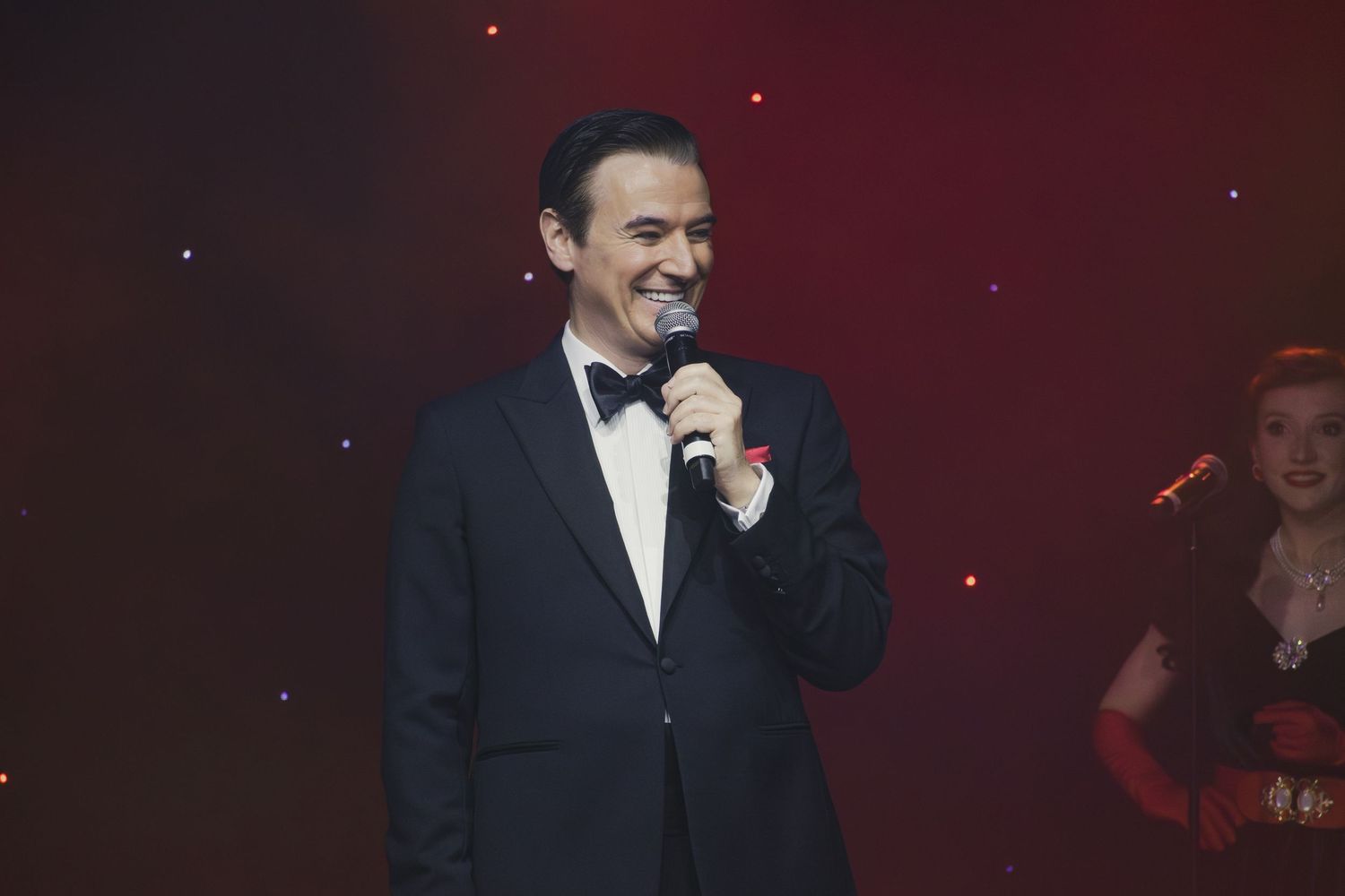 A performer in a classic tuxedo and bow tie sings on stage with dramatic red lighting.
