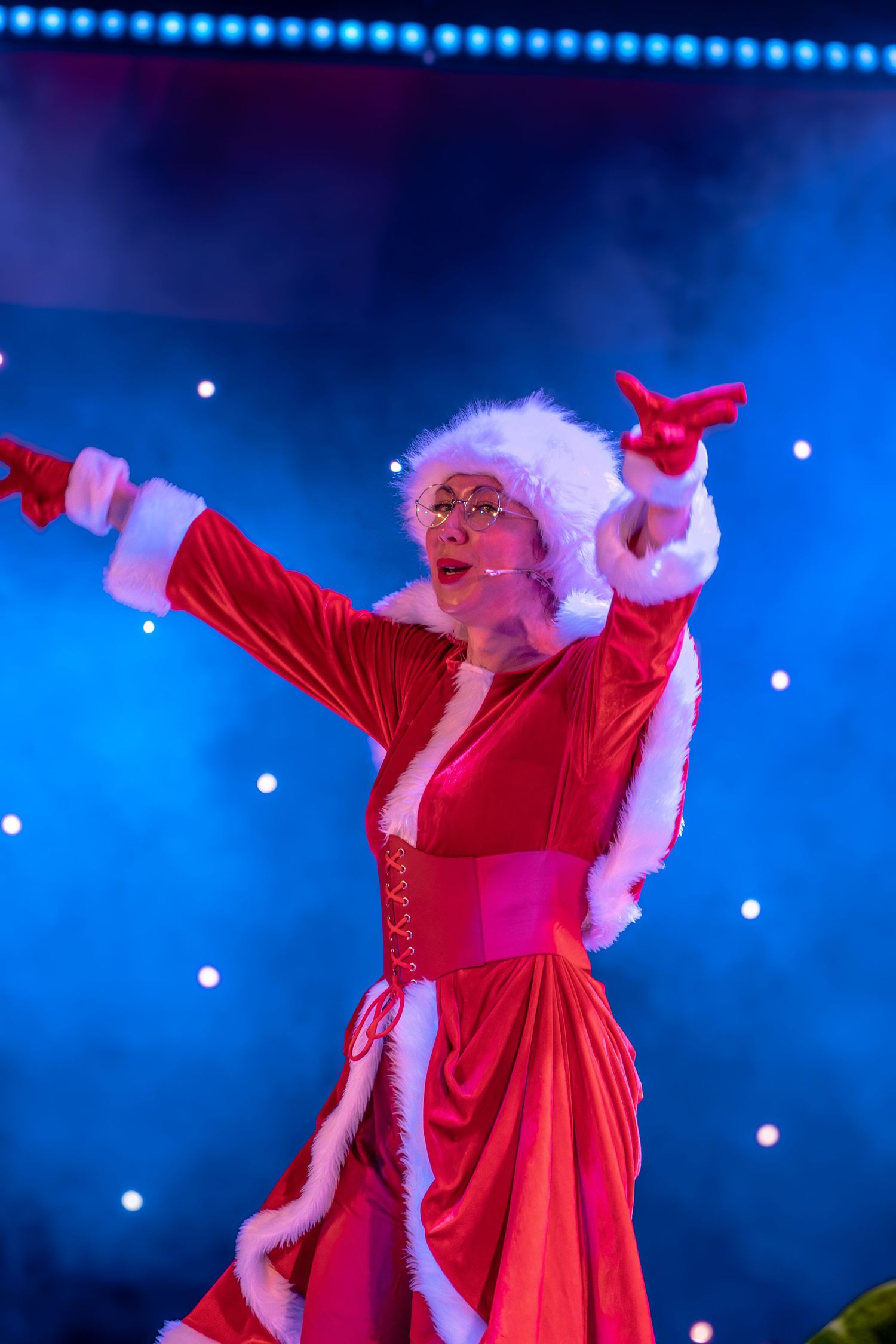 Theatrical performer in red holiday dress with arms outstretched against starlit blue background.