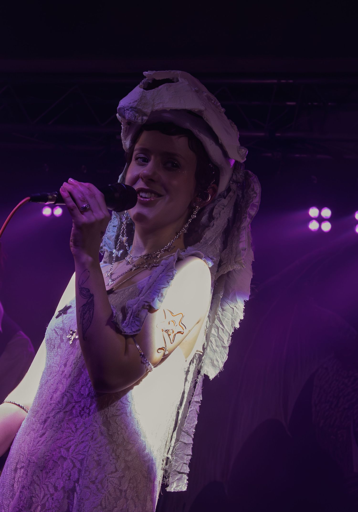 Concert performer in white outfit singing on stage under dramatic purple lighting.