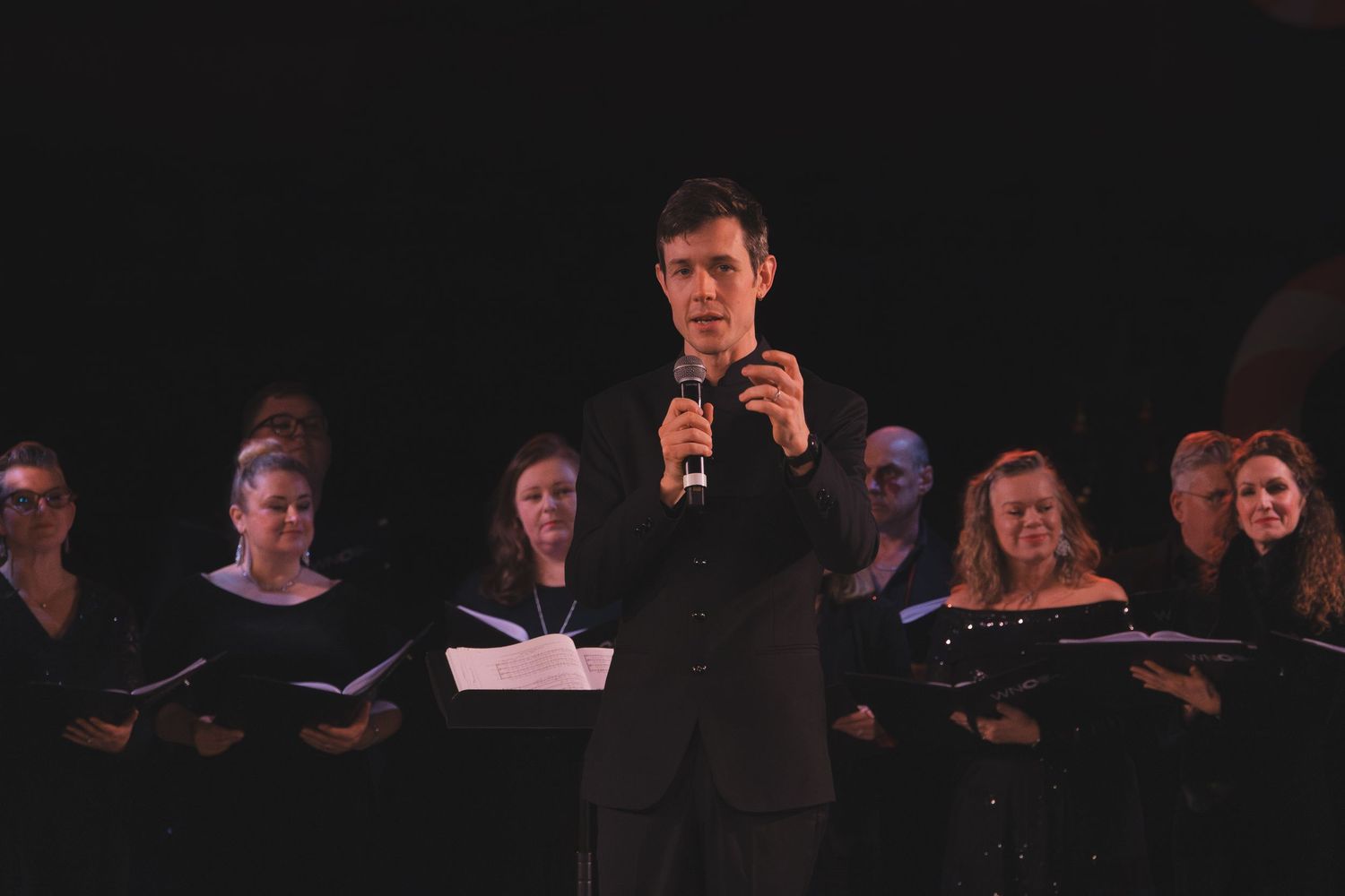 A group performs on stage in a dark theater setting with music stands and spotlights.