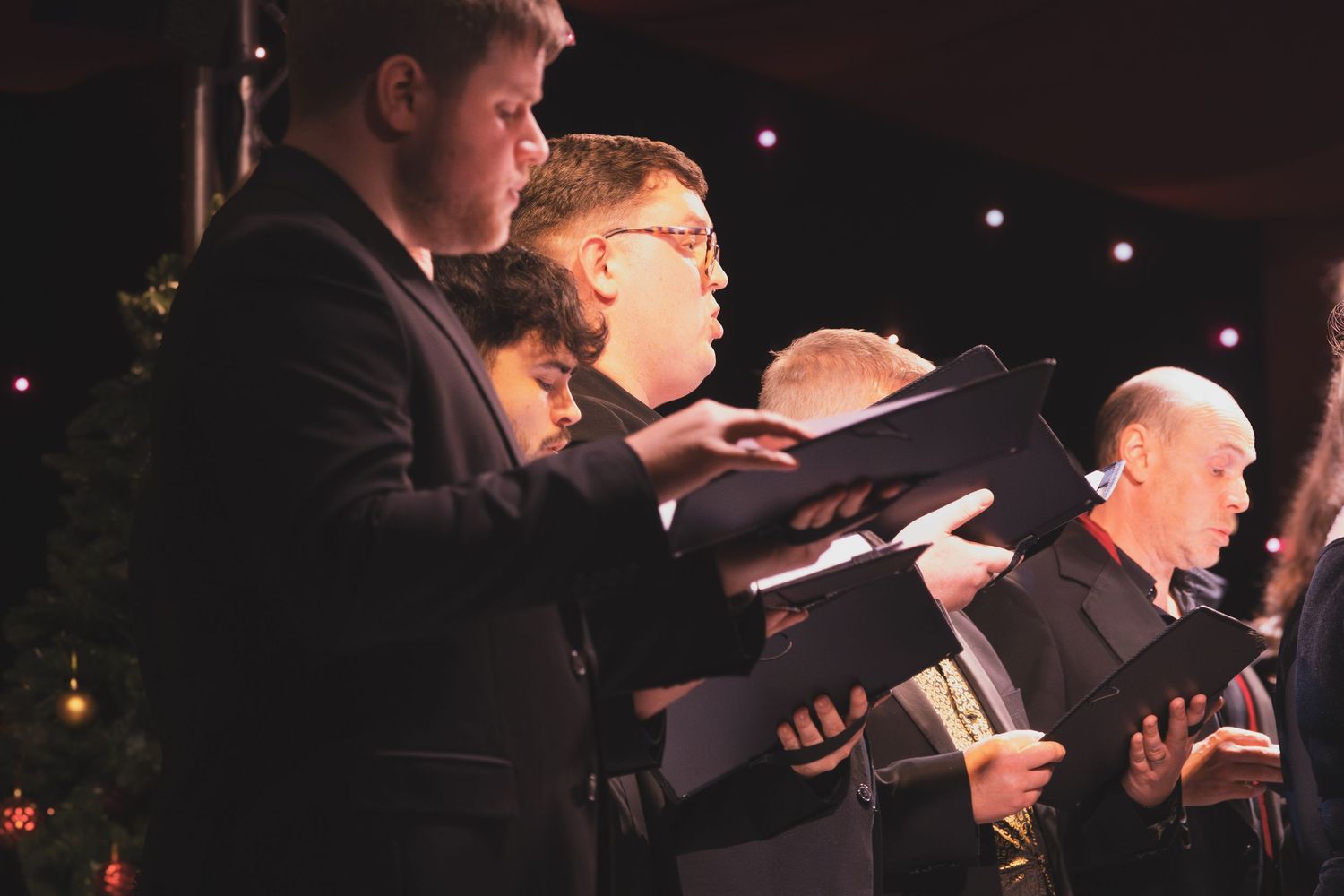 Choir performers in black attire sing together while holding sheet music during an evening performance.
