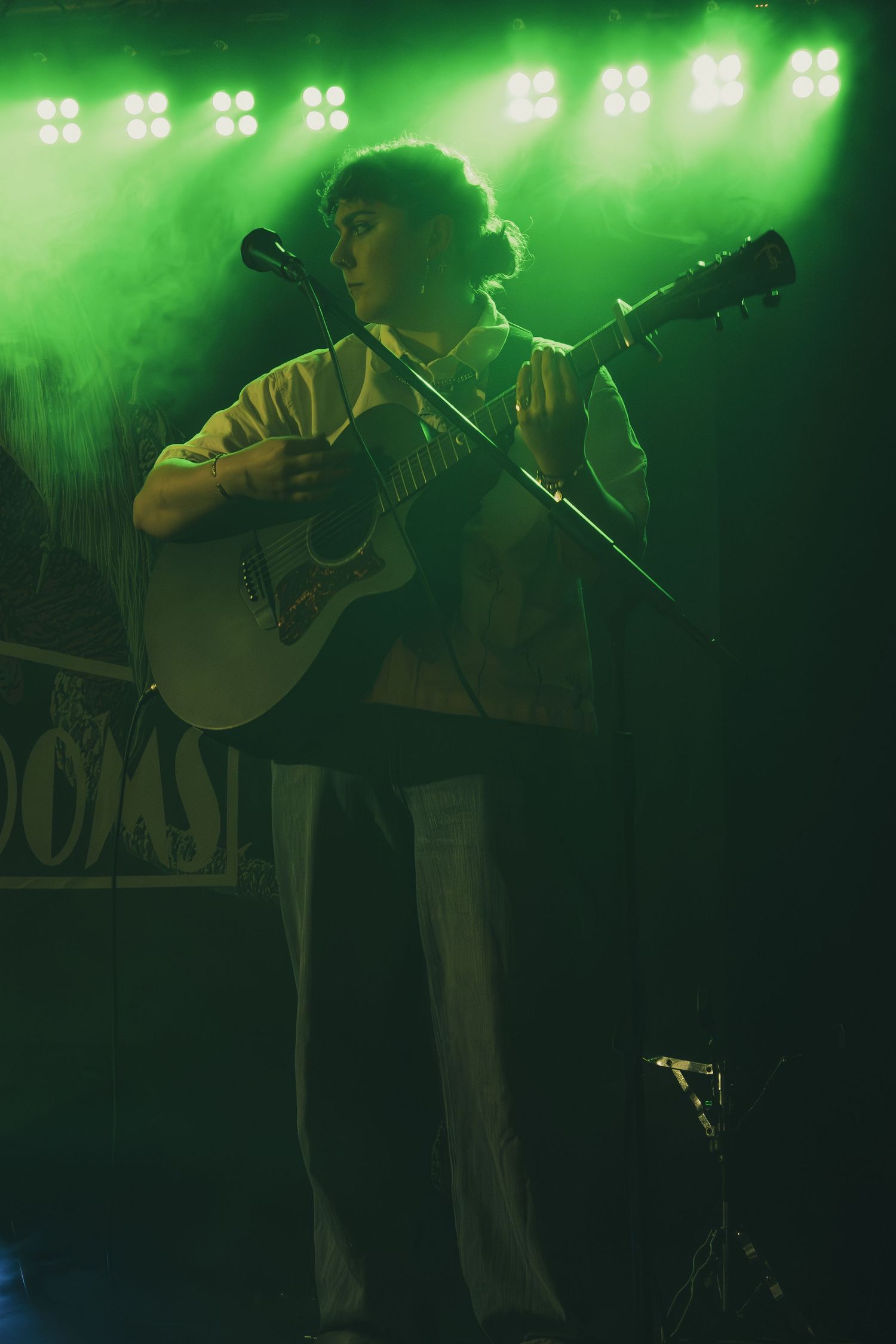 Musician performing on stage with acoustic guitar under bright green stage lighting during live concert performance.