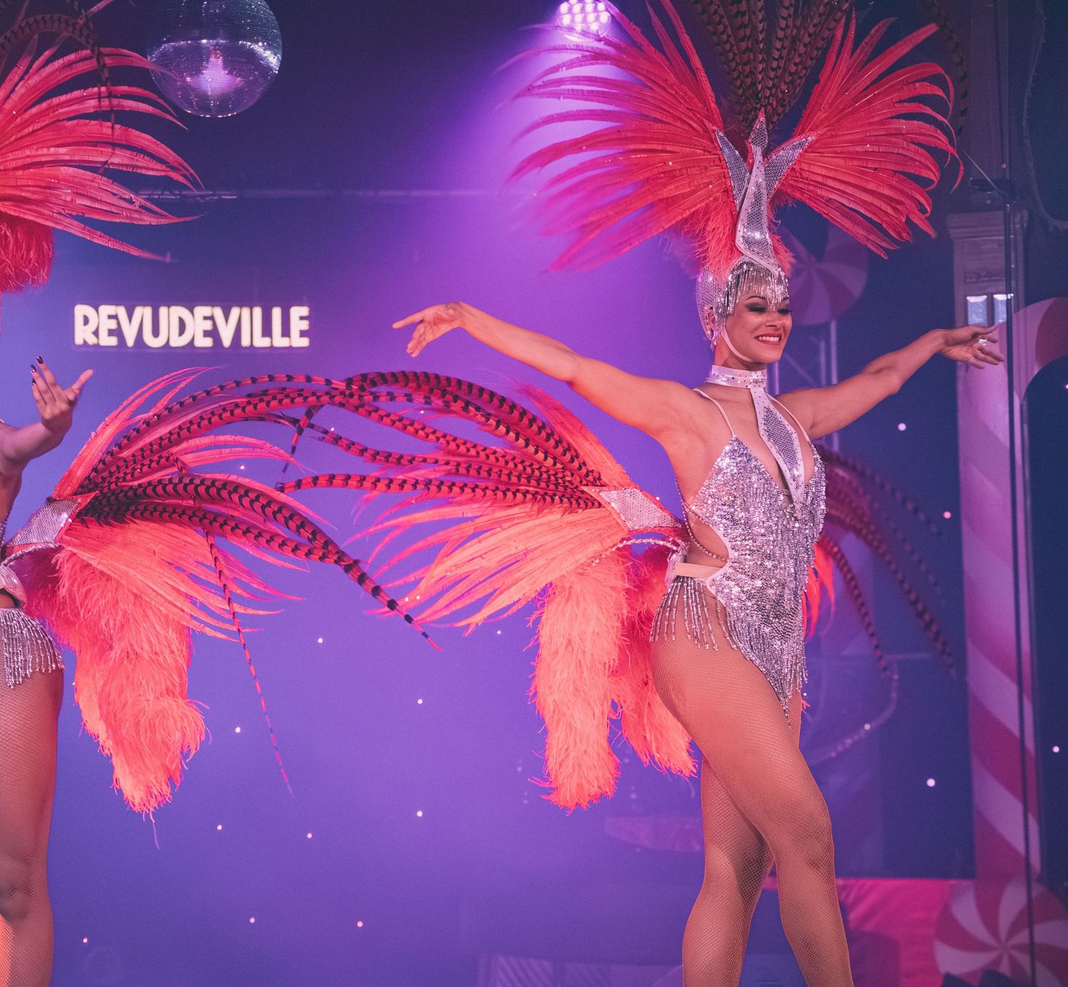 A performer in a glittering costume with bright pink feathers dances under purple stage lights.