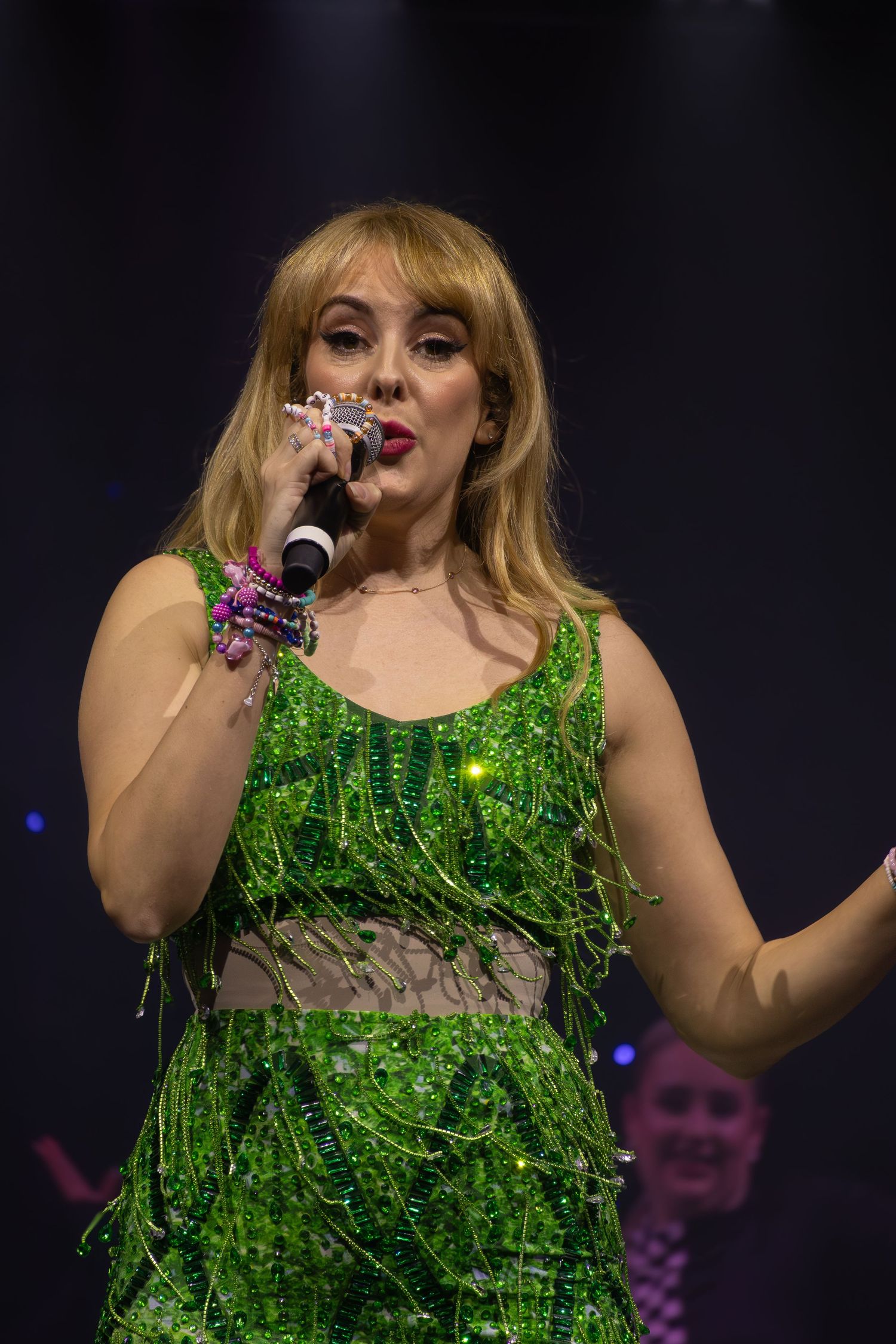 Concert performer in a sparkly green fringe dress singing on stage with dramatic lighting and dark background.