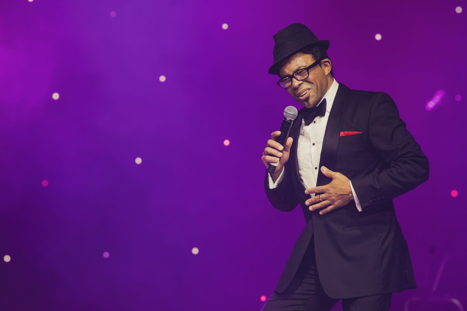 A performer in glasses and formal attire sings passionately on stage with purple lighting effects.